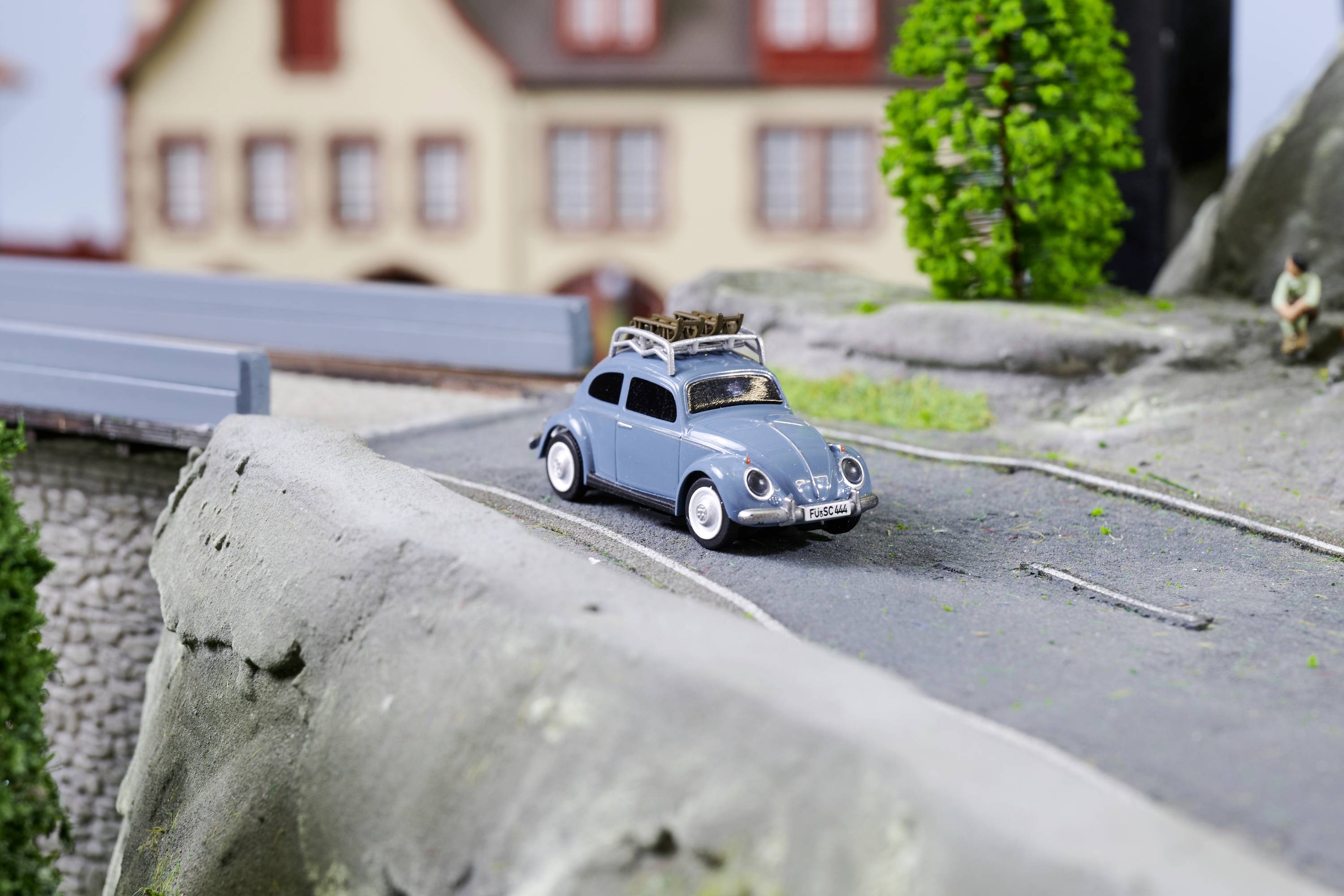 Šedé modelové auto jede po miniaturní silnici v modelované městské krajině. V pozadí stojí budovy.