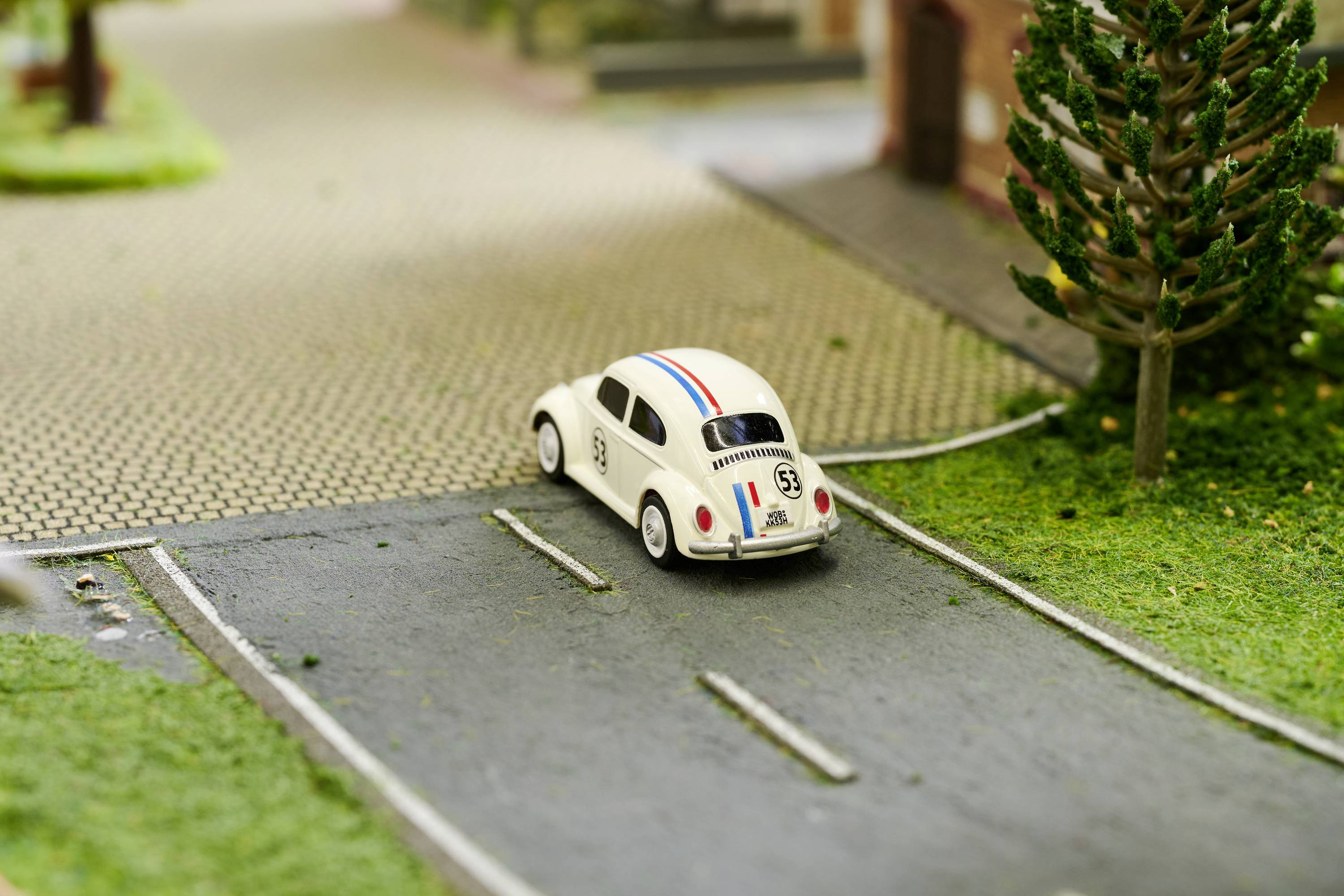 Bílé hračkové auto s racing pruhy jede po modelové silnici v miniaturní krajině s umělou trávou a stromy.