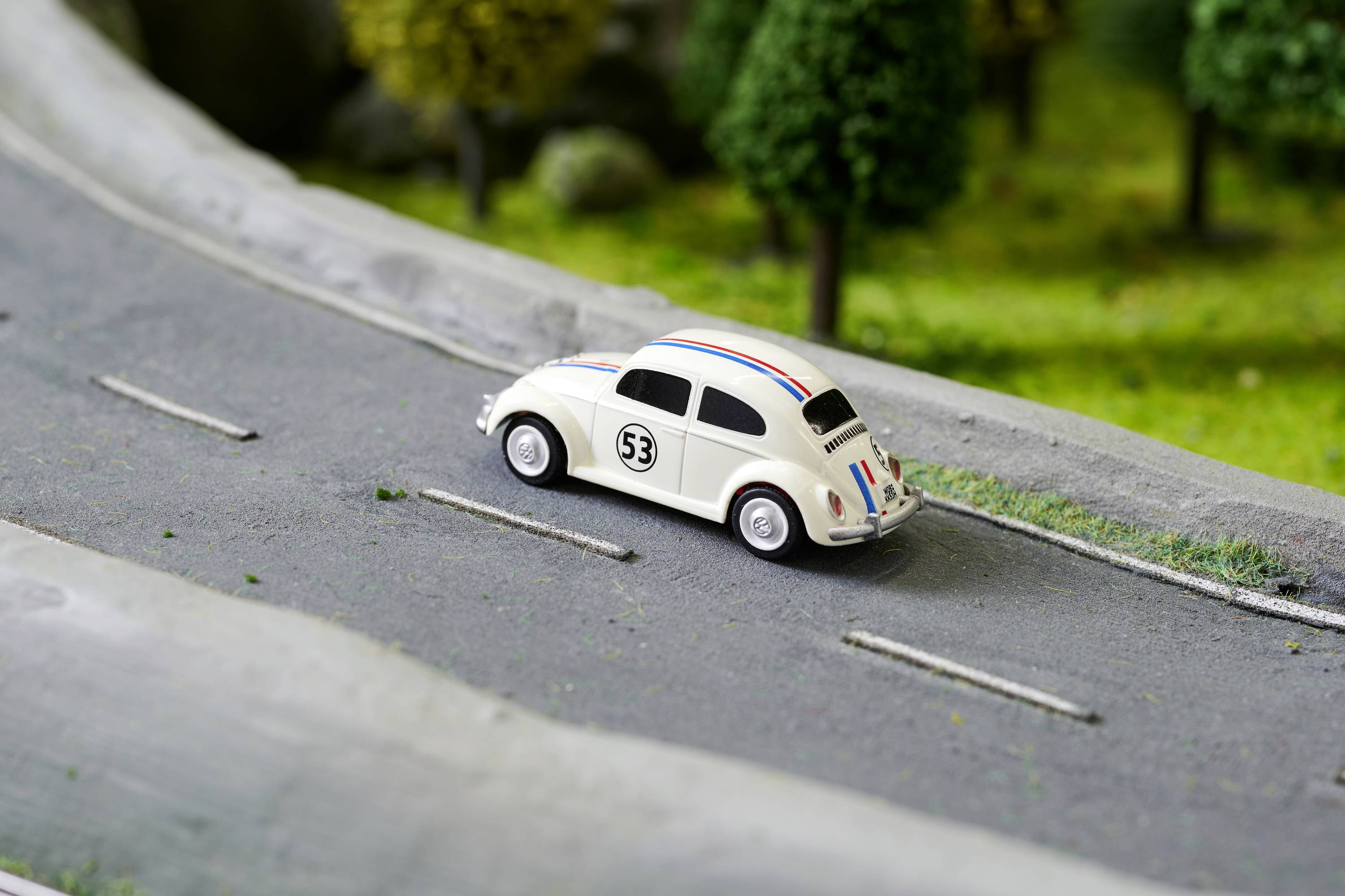Bílé hračkářské auto ve stylu klasického volkswagenu brouka jede po miniaturní silnici, obklopené zelenou modelovou trávou.