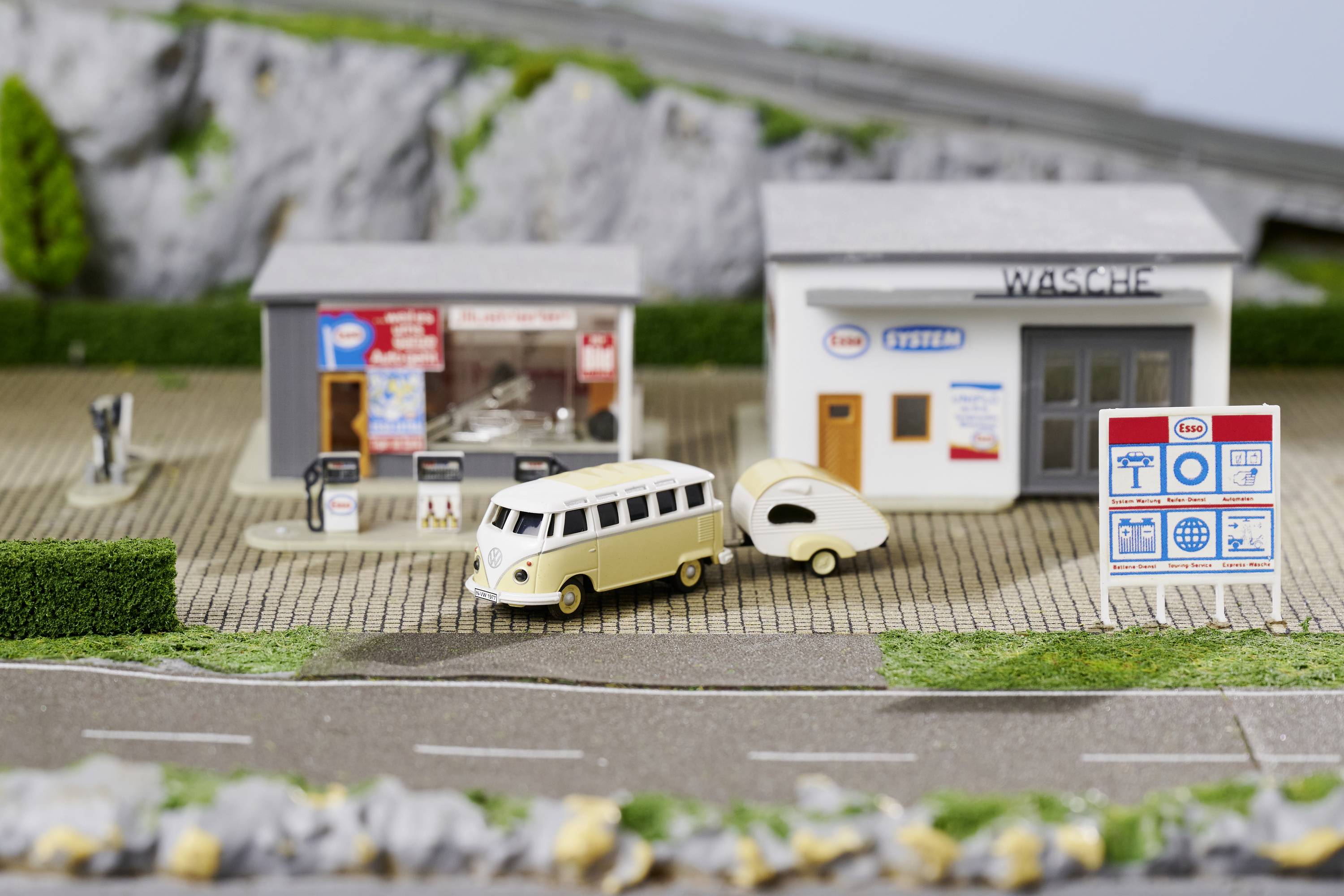 Model kempu s klasickým VW busem tažícím obytný přívěs. V pozadí jsou budovy a cedule s nápisem.
