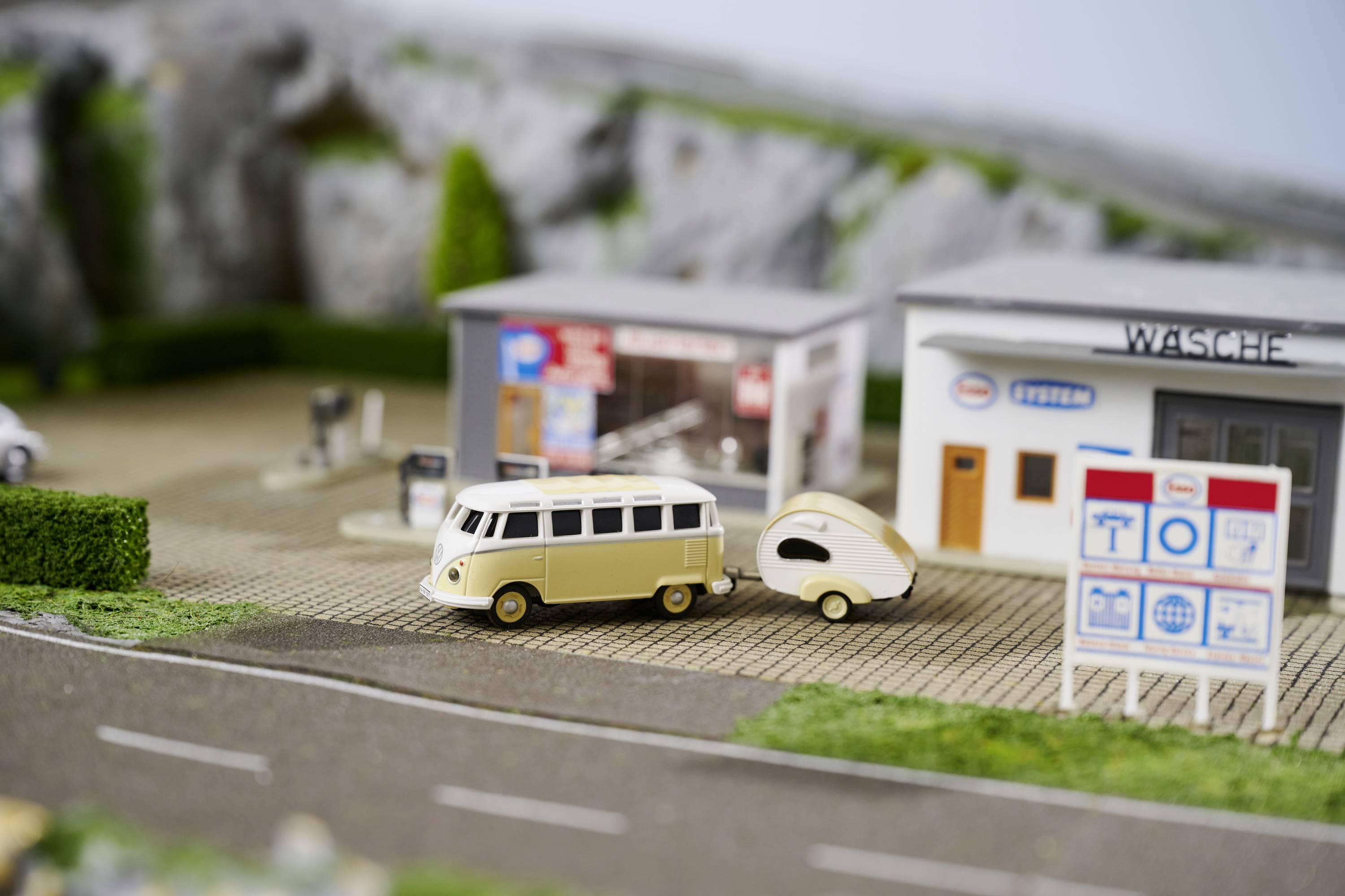 Béžové modelové auto s přívěsem stojí na dioramatu před čerpací stanicí. V pozadí jsou vidět modely budov a stromů.