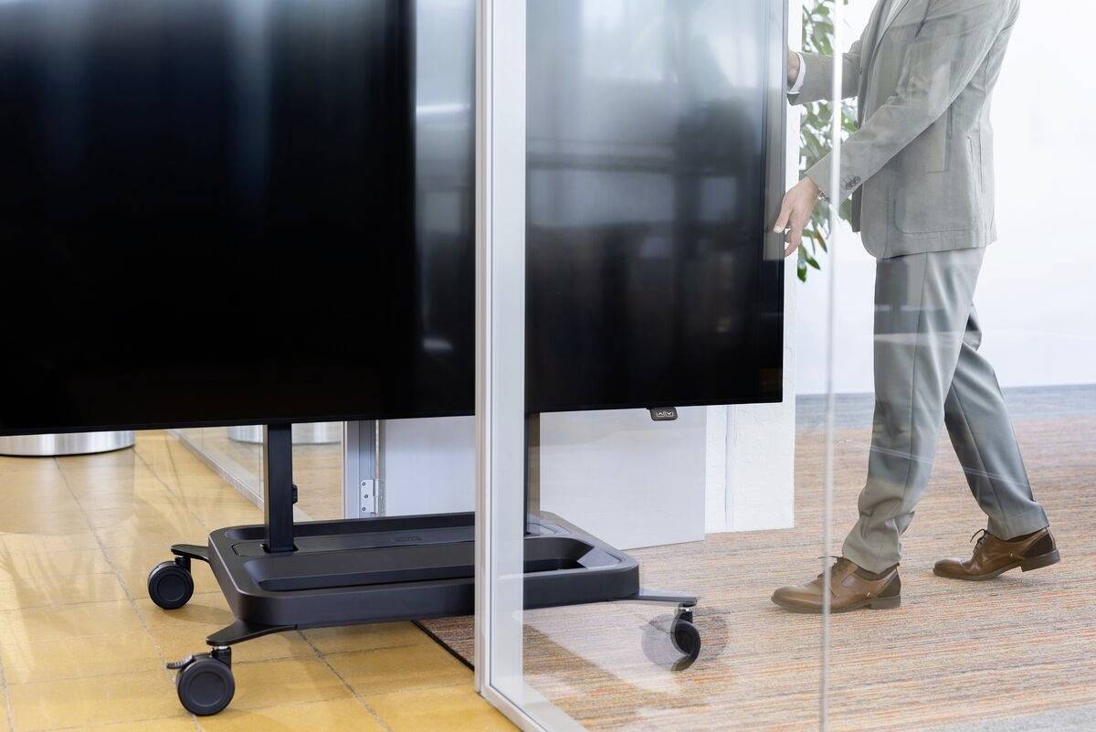 Muž v kancelářském obleku tlačí velký monitor na kolečkách skleněným průchodem. V pozadí jsou viditelné rostliny.