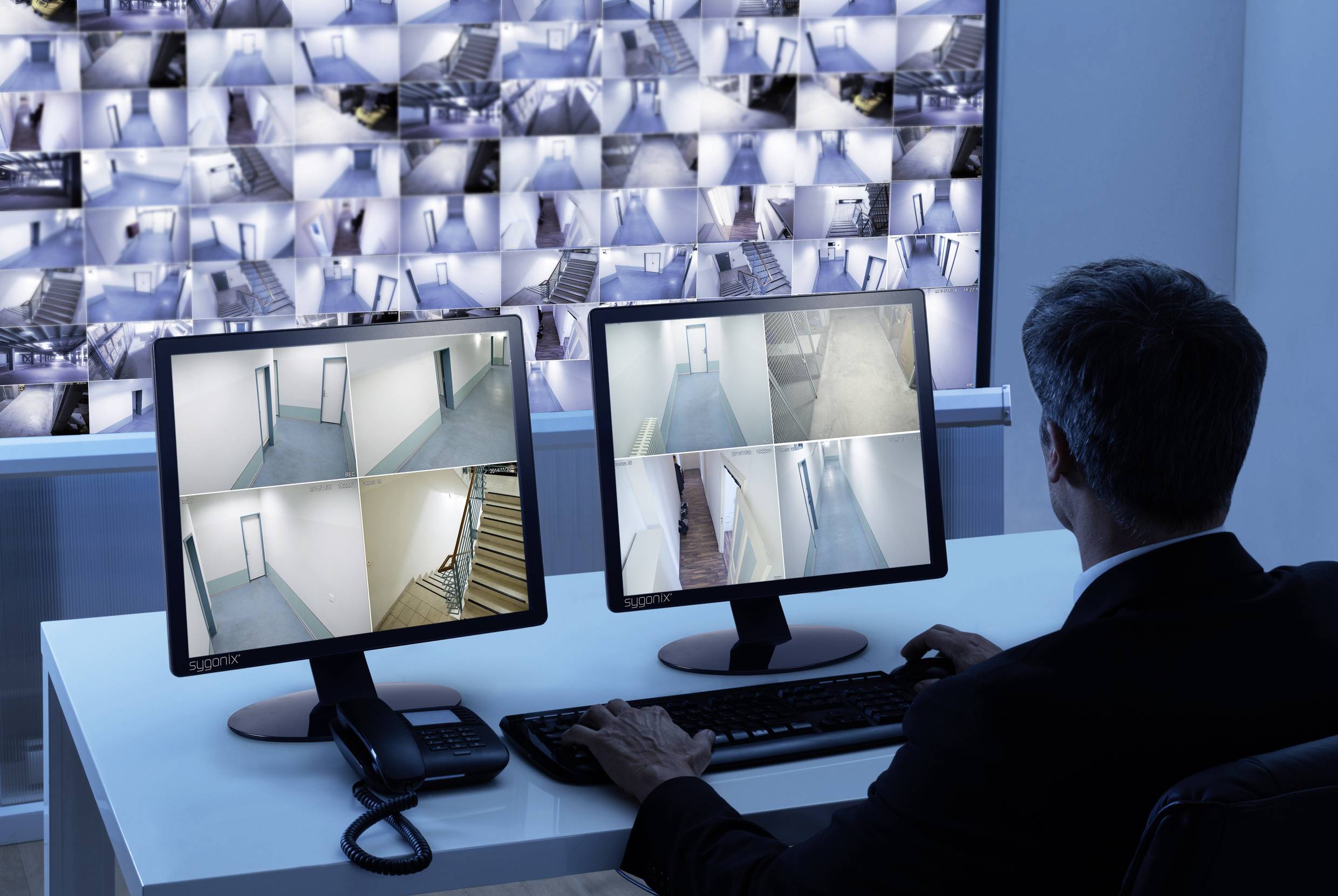 Muž monitoruje několik bezpečnostních kamer na dvou obrazovkách, které zobrazují různé části budovy. V pozadí je vidět mnoho kamerových záběrů.