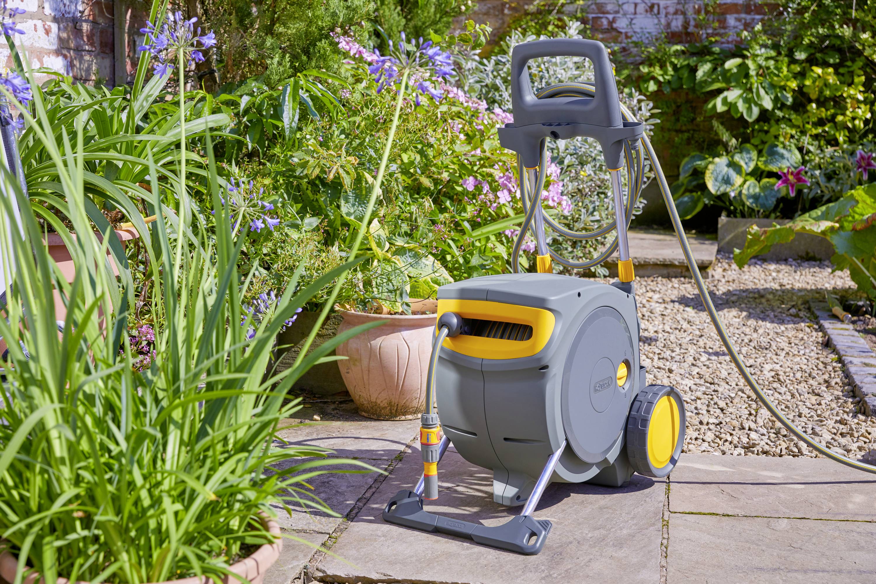 Přenosný vozík na zahradní hadici stojí na terase, obklopený zelenými rostlinami a kvetoucími květinami v květináčích.