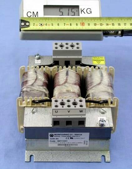 Transformátor na modré ploše, nad ním měřicí jednotka. Display ukazuje '5,75 KG'. Přes to je položeno svinovací metr.