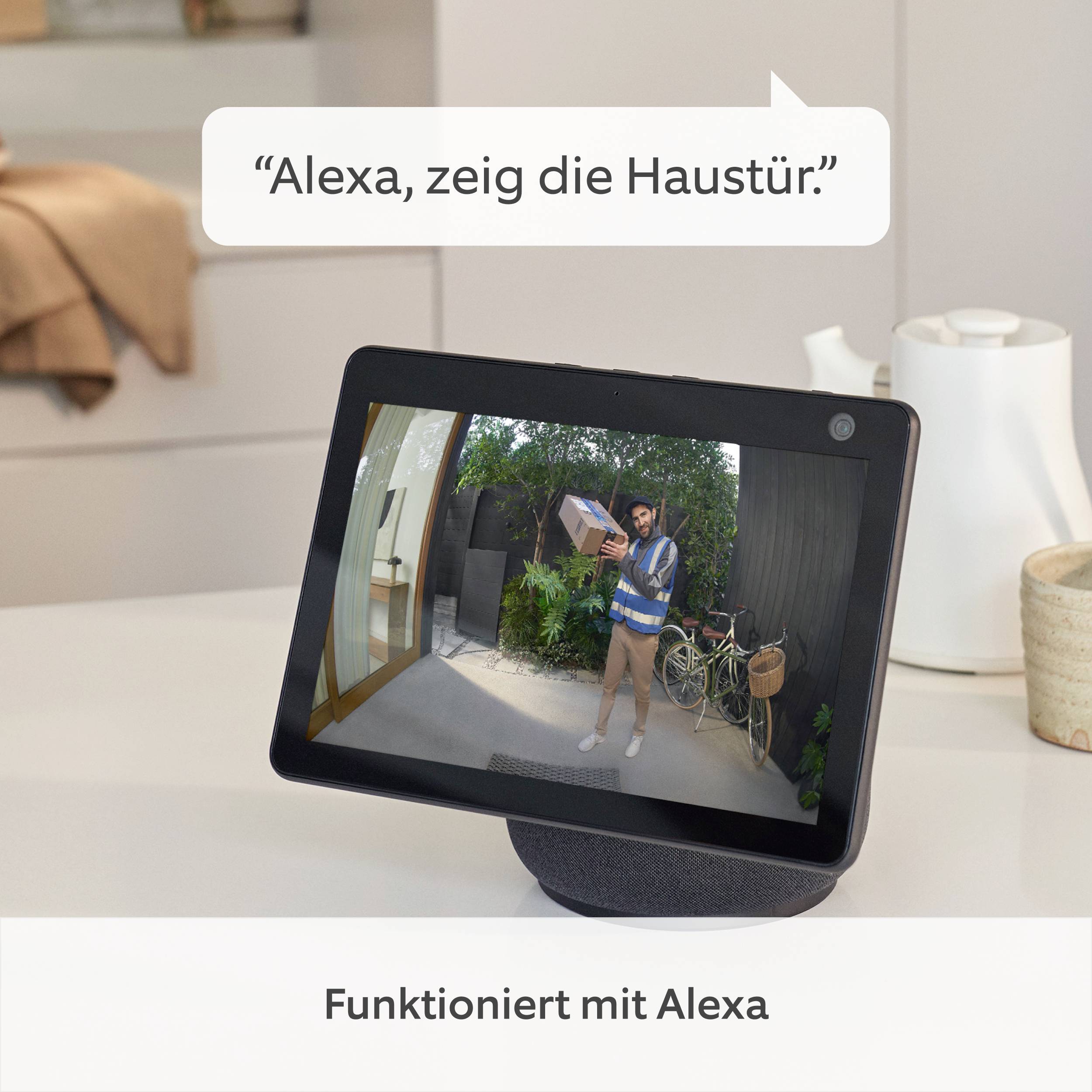 Obrazovka zobrazuje doručovatele balíku před dveřmi. Nahoře stojí 'Alexa, ukaž vstupní dveře.' Dole: 'Funguje s Alexou'.