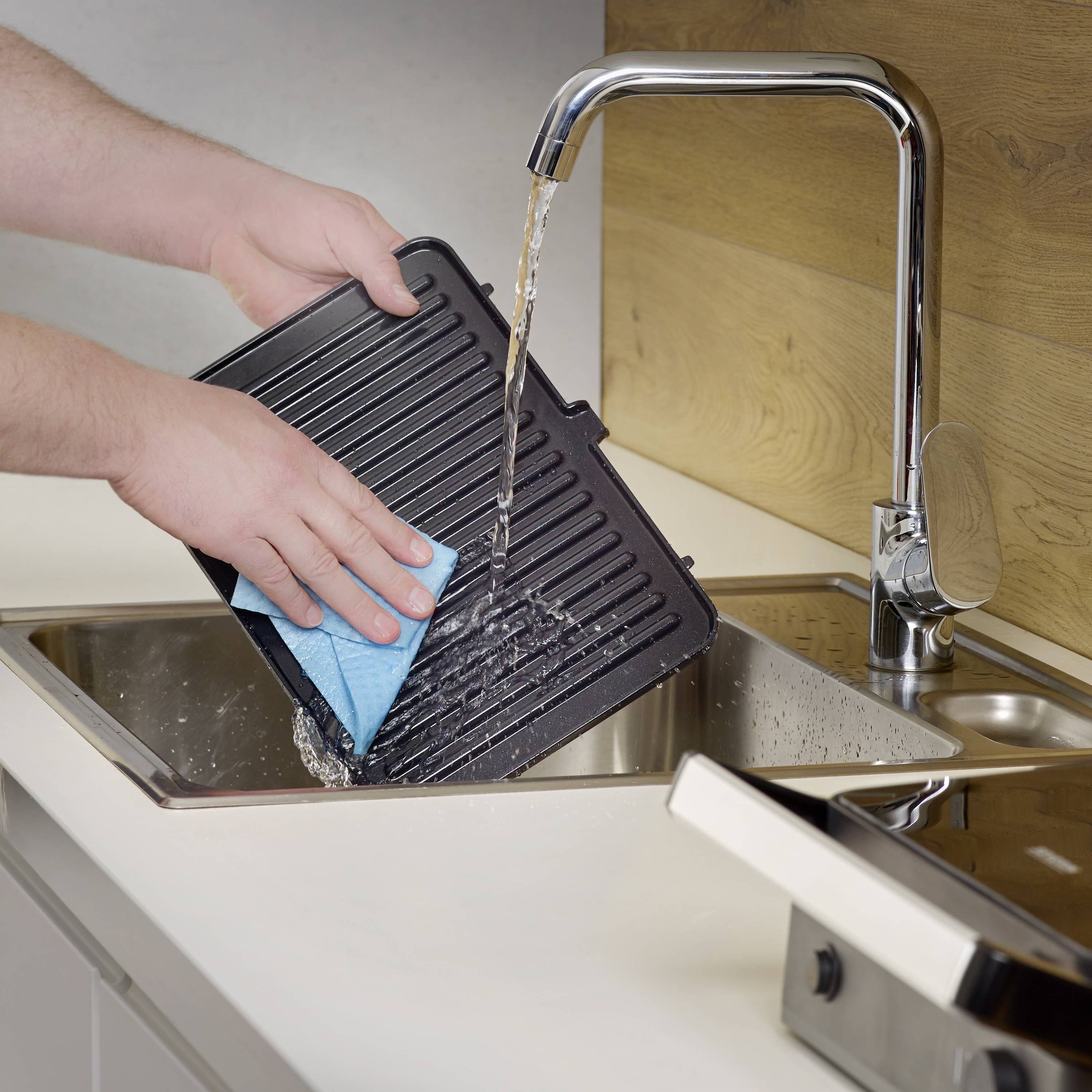 Osoba oplachuje černý grilovací plech vodou a modrým houbičkou v nerezovém dřezu.