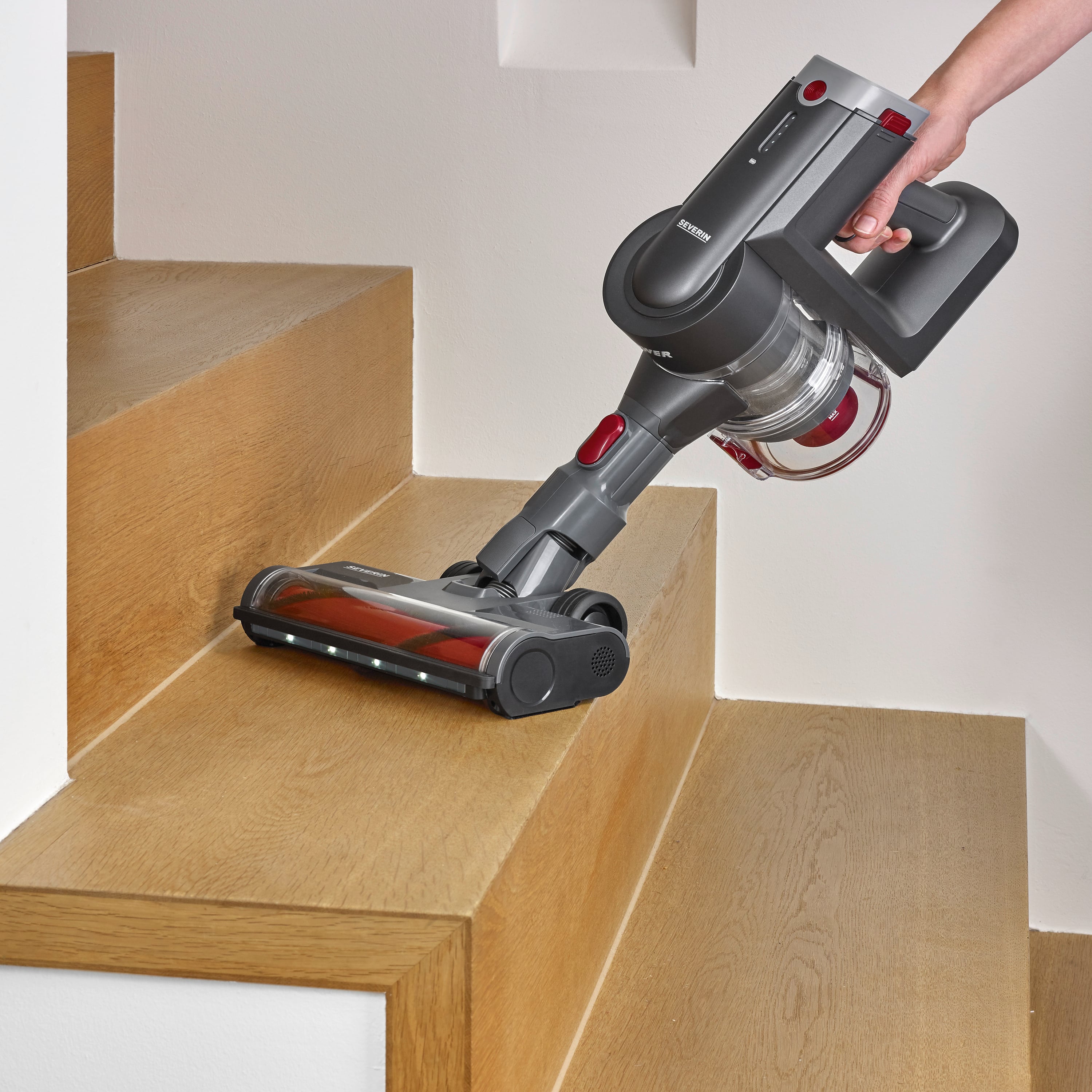 Bezdrátový vysavač čistí dřevěné schody. Jedna ruka drží rukověť vysavače, zatímco kartáč přejíždí přes stupně.