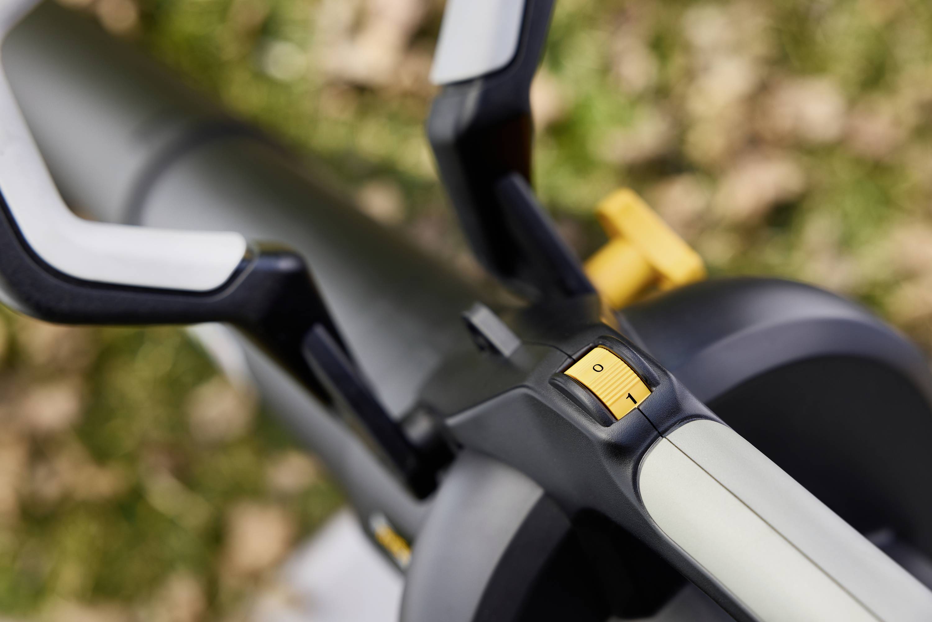 Detailní záběr elektrické foukače listí se spínačem zapnuto/vypnuto. V pozadí rozostřené listí na trávníku.
