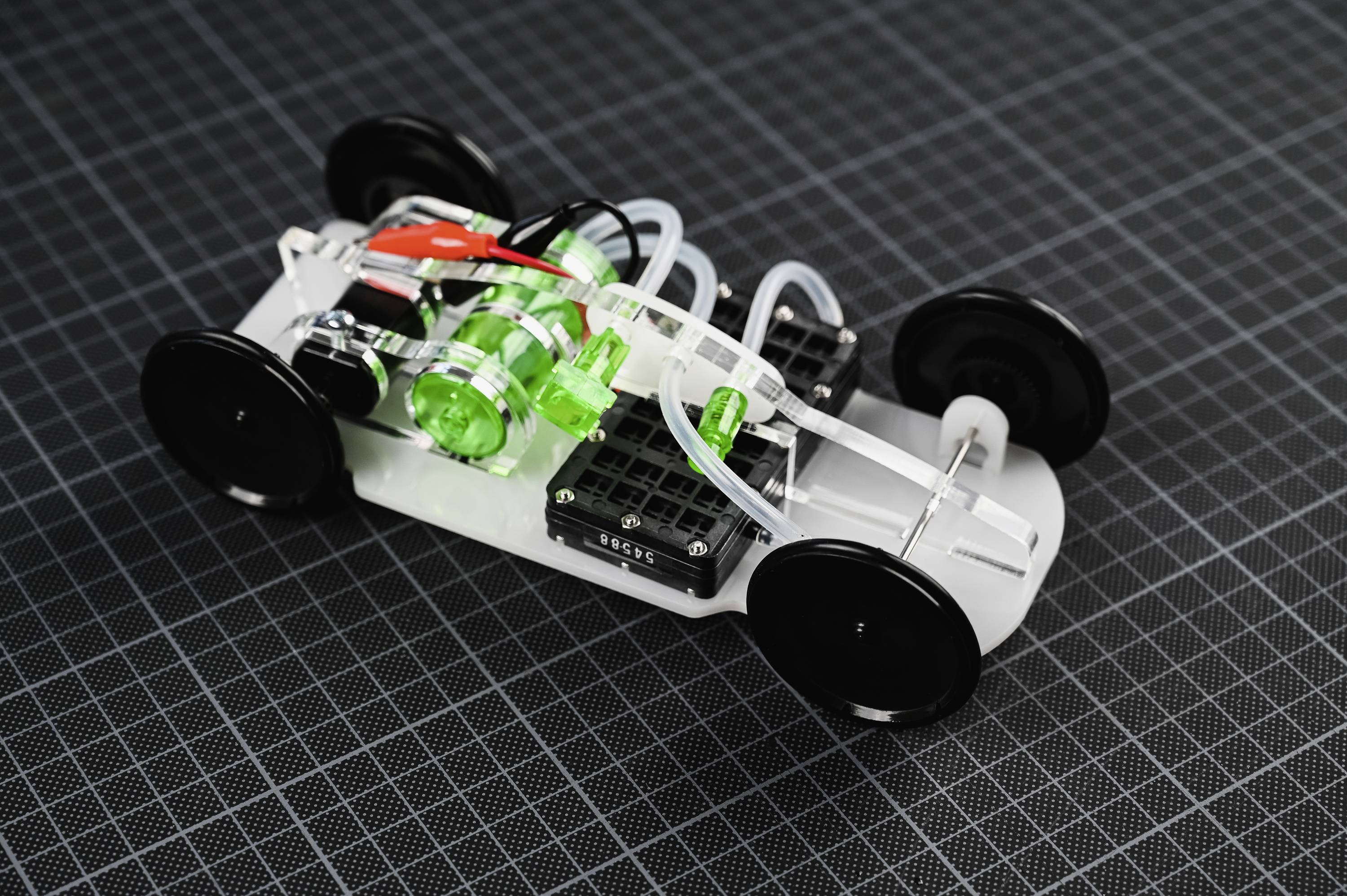 Malý, průhledný modelový vůz s zelenými a černými díly na kostkovaném pozadí ukazuje technický a robotický design.