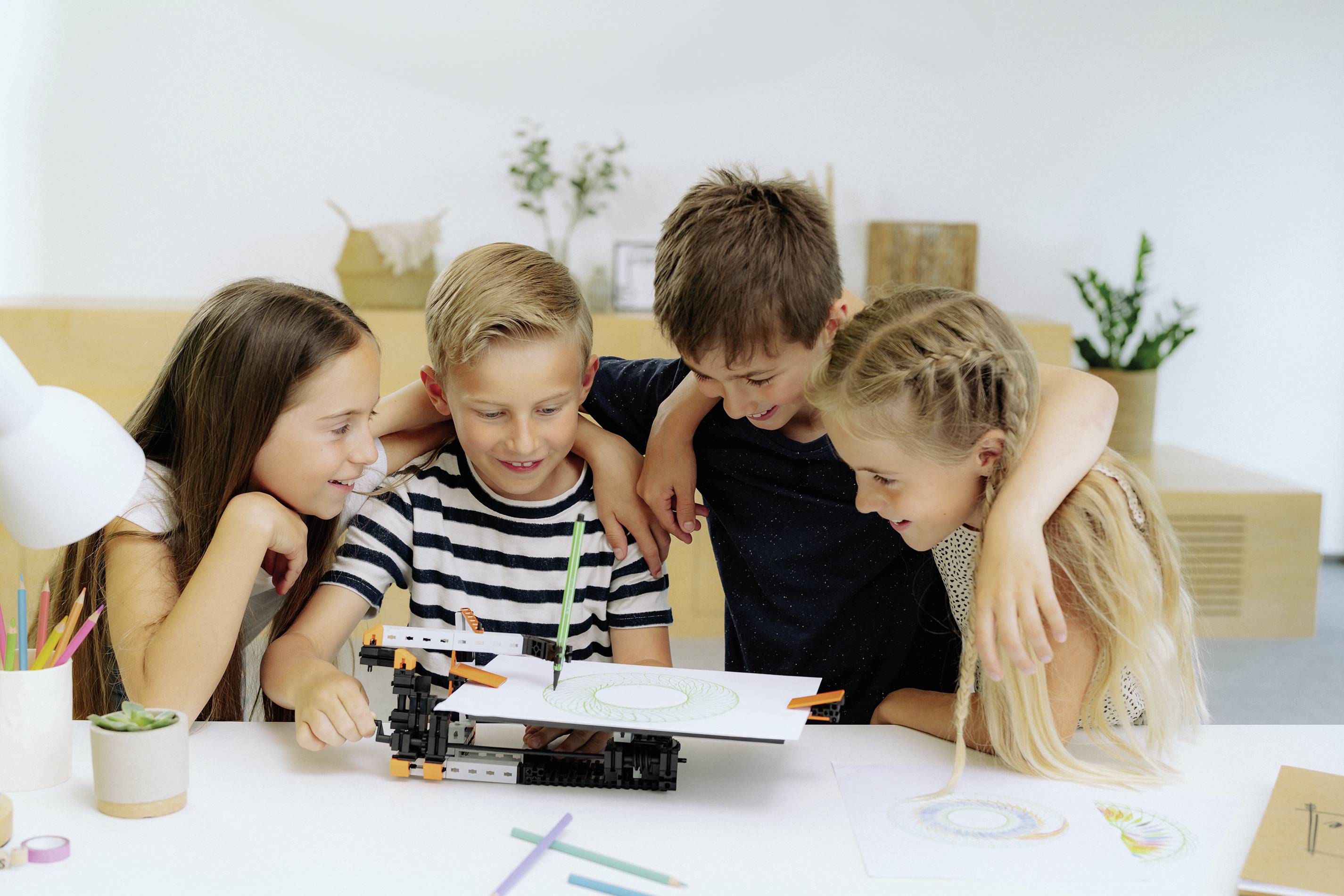 Čtyři děti sedí společně u stolu a prohlížejí si projekt z papíru a stavebnicových dílů. Vypadají soustředěně a vesele.