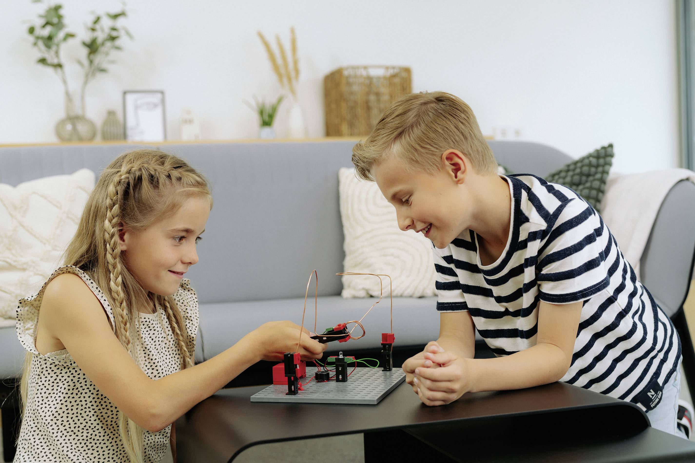Dívka a chlapec sedí u stolu a experimentují s malým elektronickým obvodem. Oba působí soustředěným dojmem.