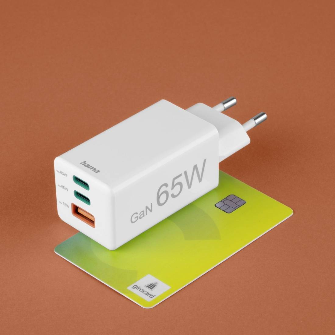 Bílá 65W GaN nabíječka od značky 'hama' se dvěma USB-C a jedním USB-A portem leží na zelené kartě na oranžovém pozadí.
