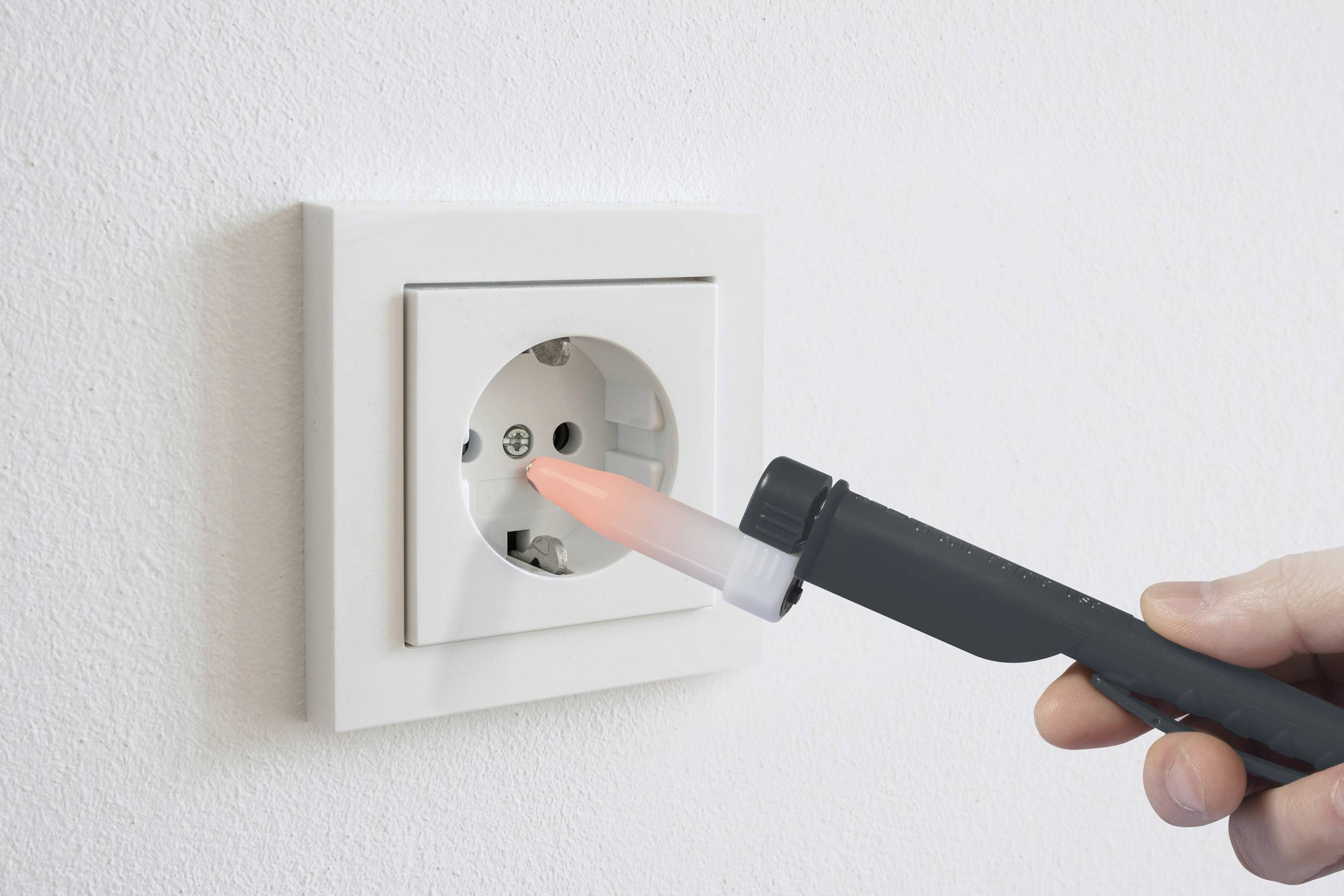 Jedna ruka drží zkoušečku napětí, jejíž špička svítí červeně, před zásuvkou, což naznačuje průtok elektrického proudu.
