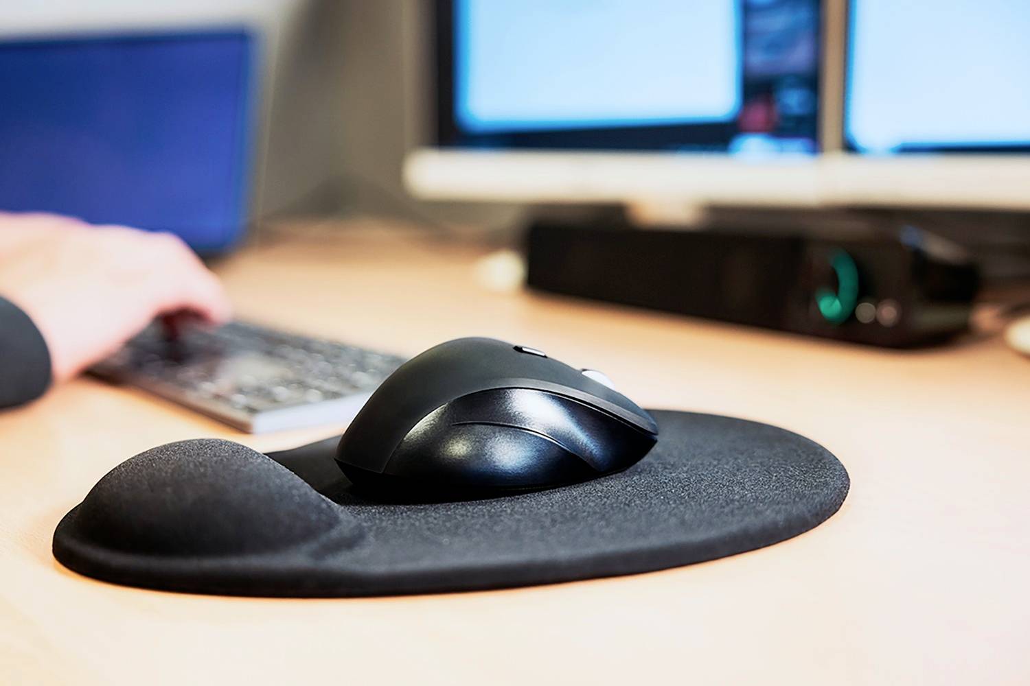 Počítačová myš a ergonomická podložka pod myš na psacím stole. V pozadí je vidět klávesnice a monitor.