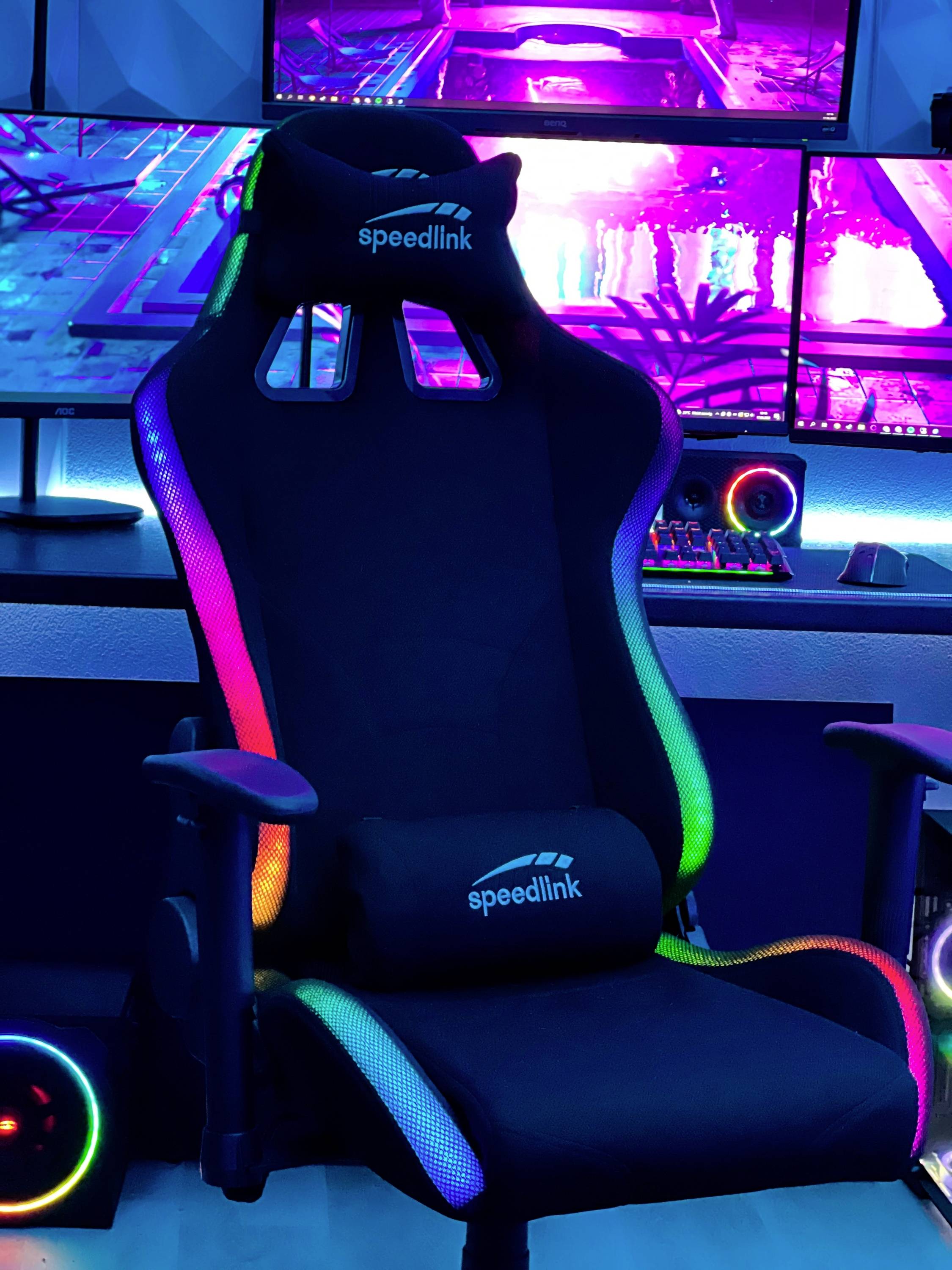Herní židle s RGB podsvícením v moderním herním prostředí. V pozadí několik monitorů s barevnými grafikami.