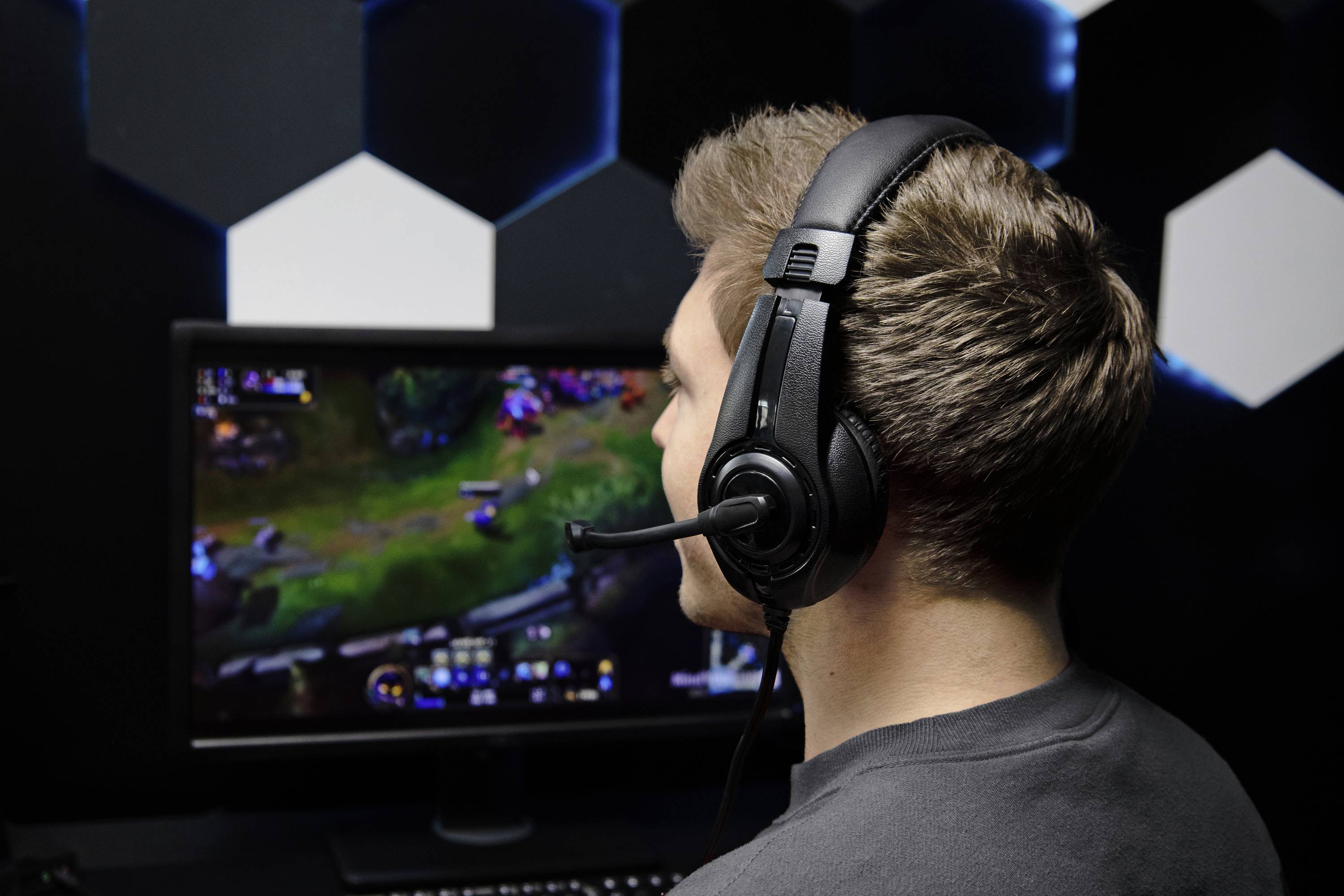 Muž se sluchátky sedí před monitorem, na kterém běží videohra. Místnost je tmavá s osvětlenými stěnami.