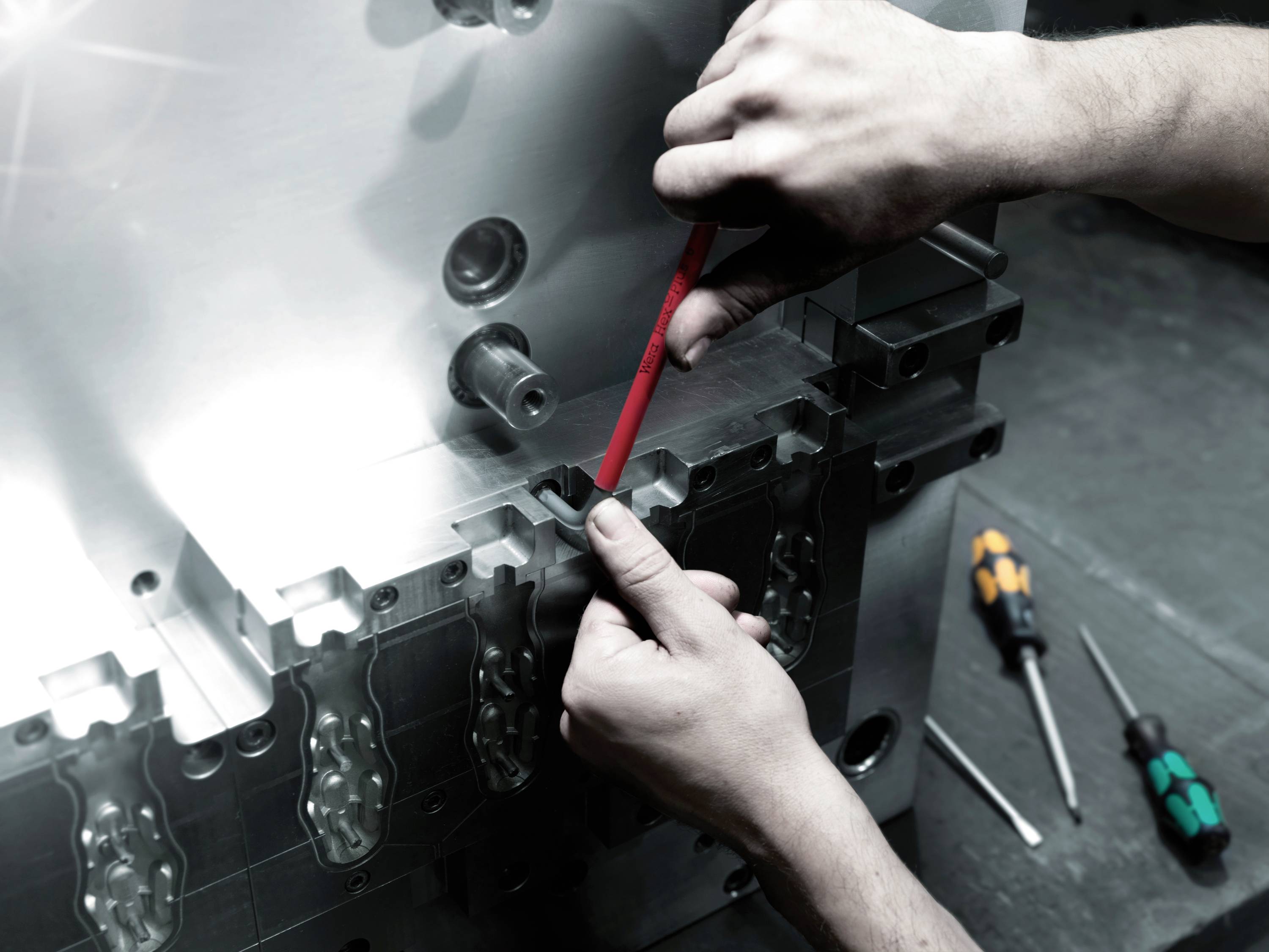 Ruce osoby používající nástroj na velké kovové strojní součásti, s šroubováky v pozadí.