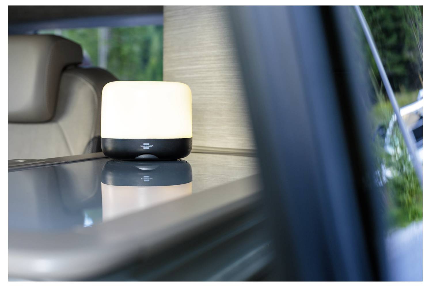 Malá svítící stolní lampa na reflexní ploše uvnitř vozidla, viděná skrz okno se sedadly v pozadí.