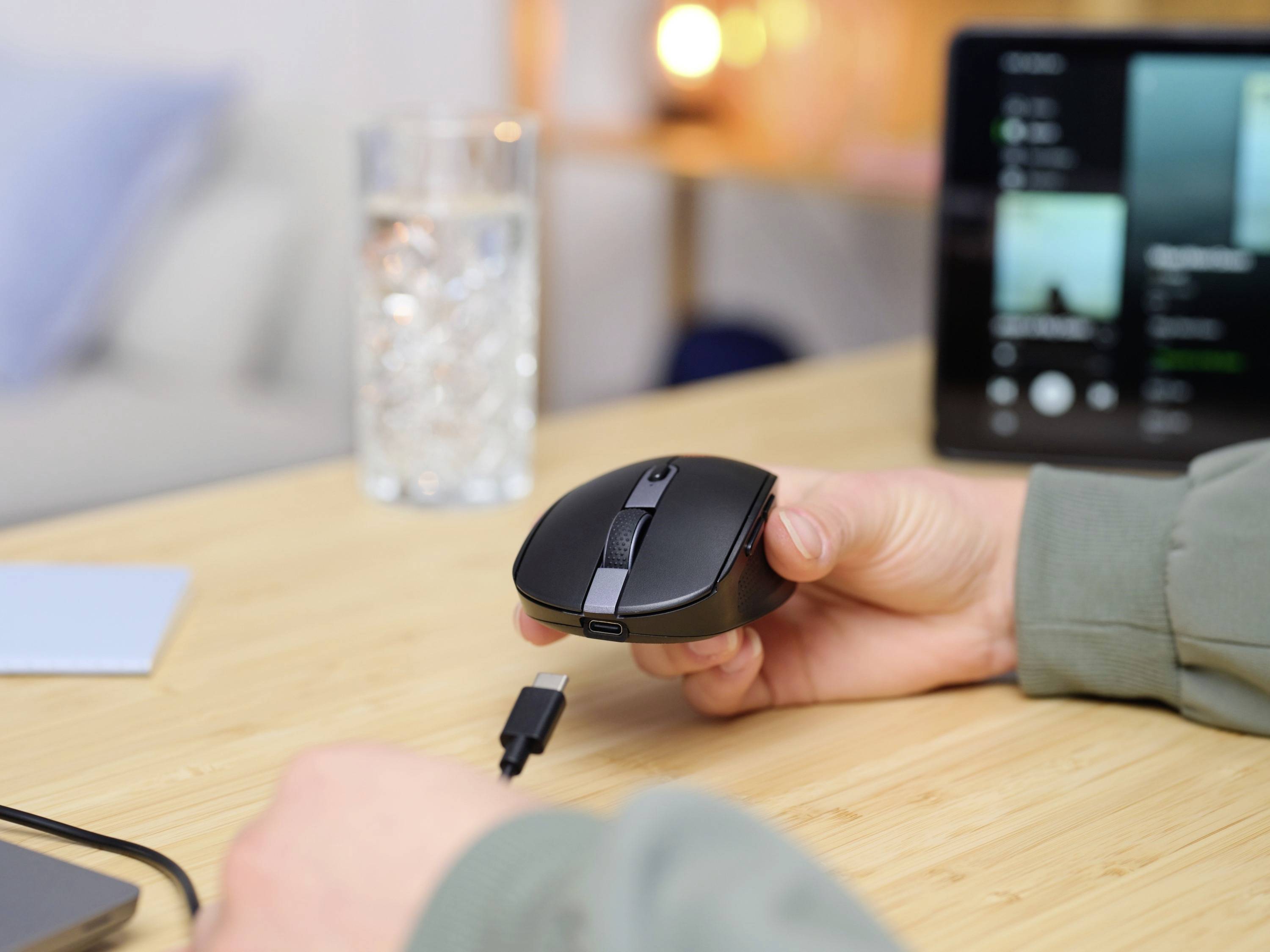 Osoba drží černou počítačovou myš, připravena připojit USB kabel. V pozadí je sklenice vody a monitor.