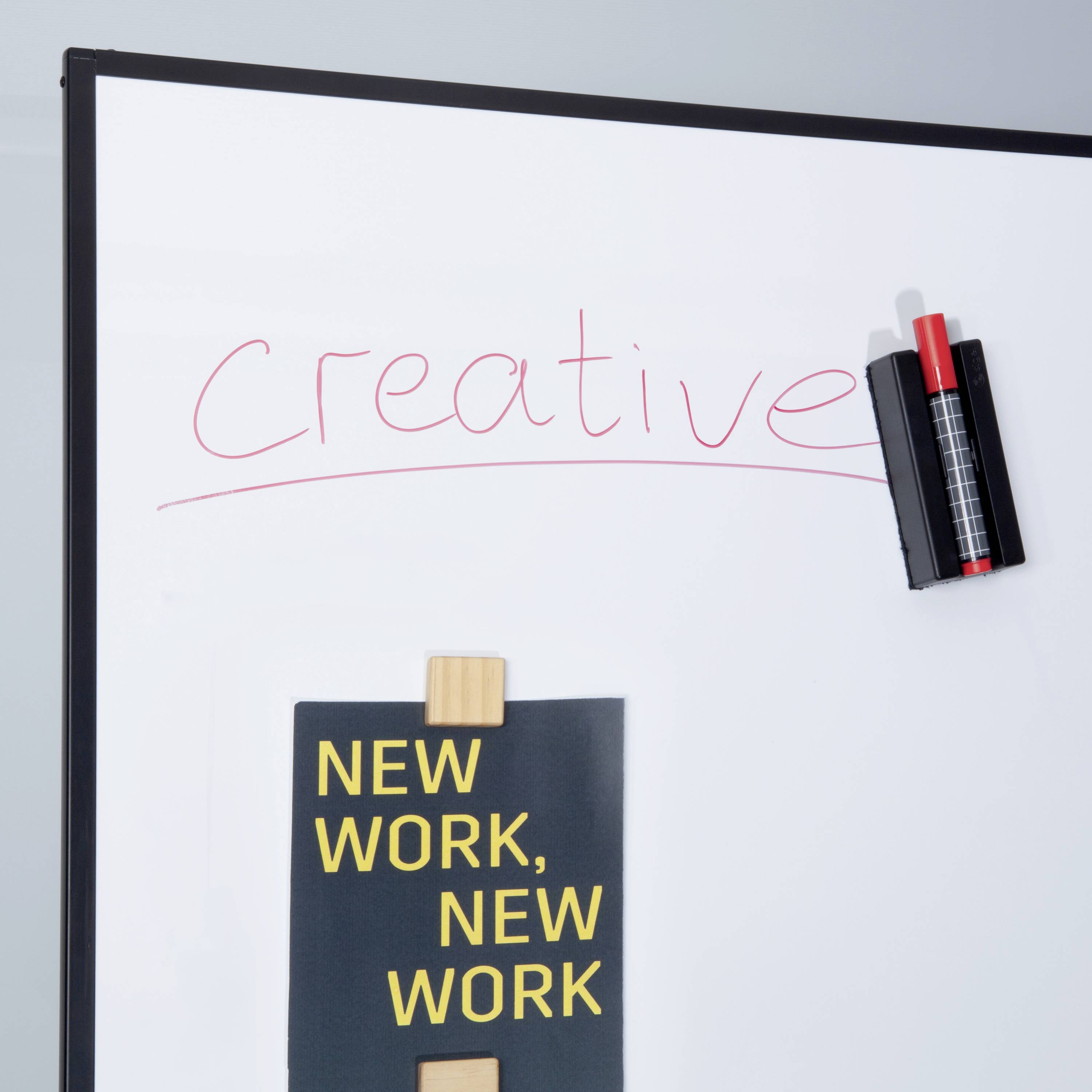 Whiteboard s nápisem 'Creative'. Pod ním malý plakát s textem 'New Work, New Work' a stojánek na psací potřeby v pravém horním rohu.