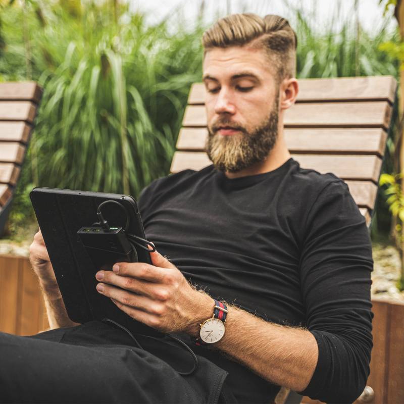 Muž s bradkou odpočívá uvolněně na lehátku a čte si na tabletu. V pozadí jsou stromy a rostliny.