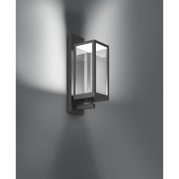 Moderní nástěnná lampa s obdélníkovým skleněným stínidlem a černým kovovým rámem, svítí proti šedé stěně a vytváří světelné efekty.