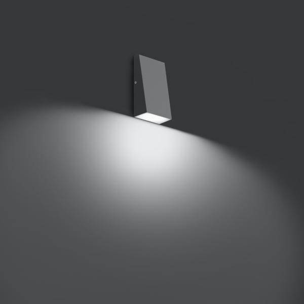 Nástěnná lampa na tmavě šedé stěně, která vyzařuje světlo dolů a vytváří jemný světelný odraz na povrchu stěny.