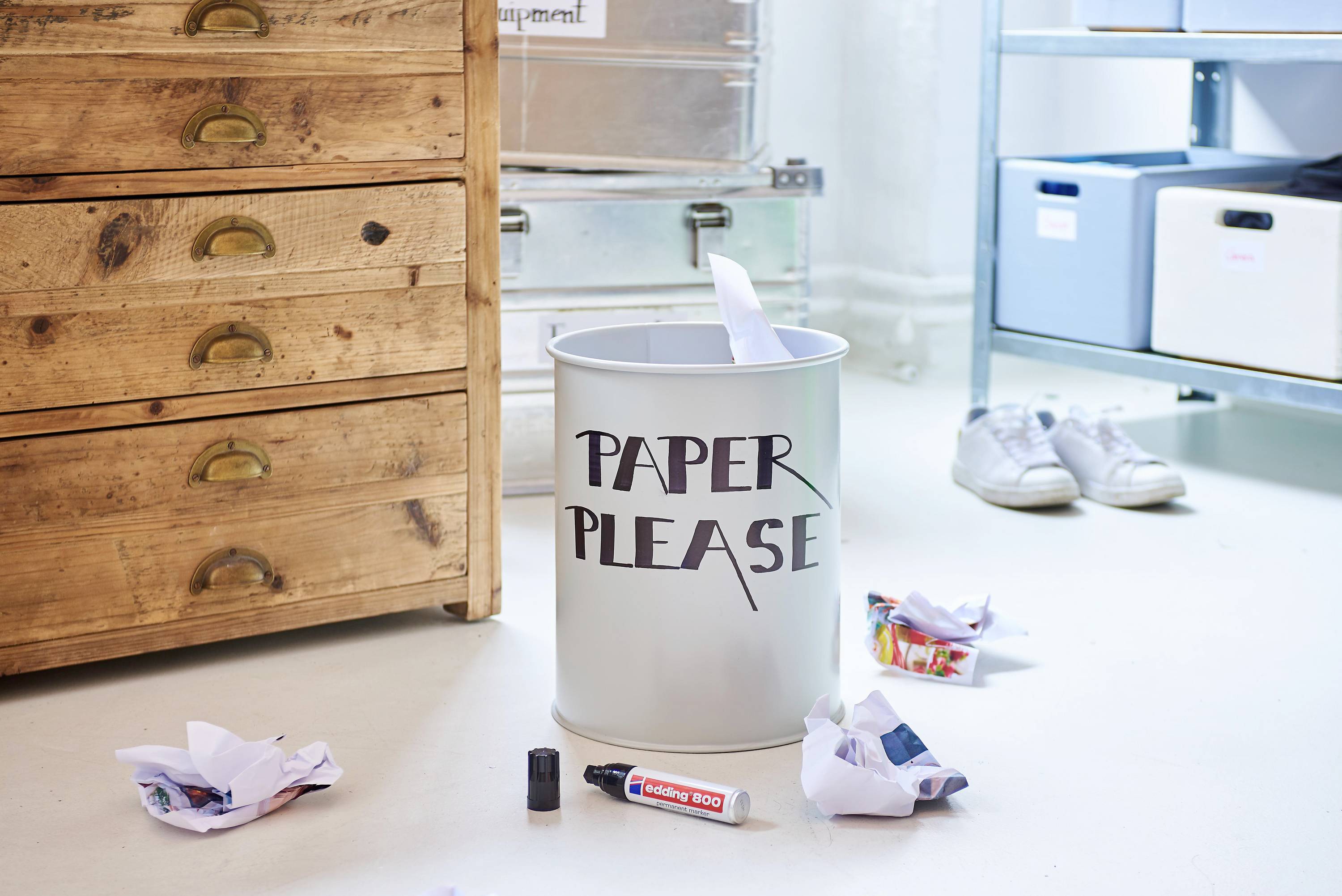 Odpadkový koš s nápisem 'Paper Please', obklopený zmačkanými papíry a kancelářskými potřebami na podlaze.
