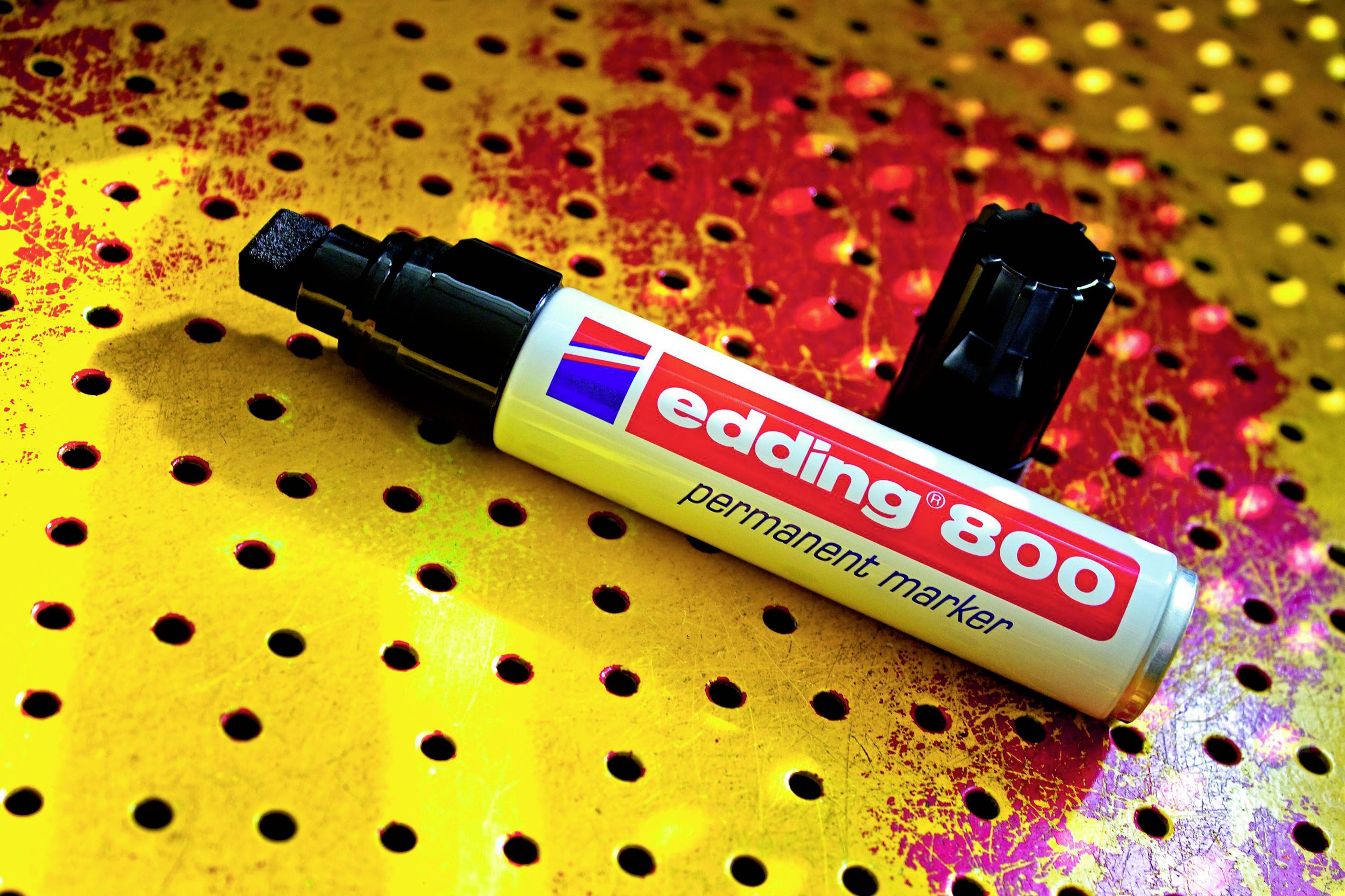 Bílý permanentní fix 'edding 800' leží otevřený na barevně posetém, děrovaném povrchu.