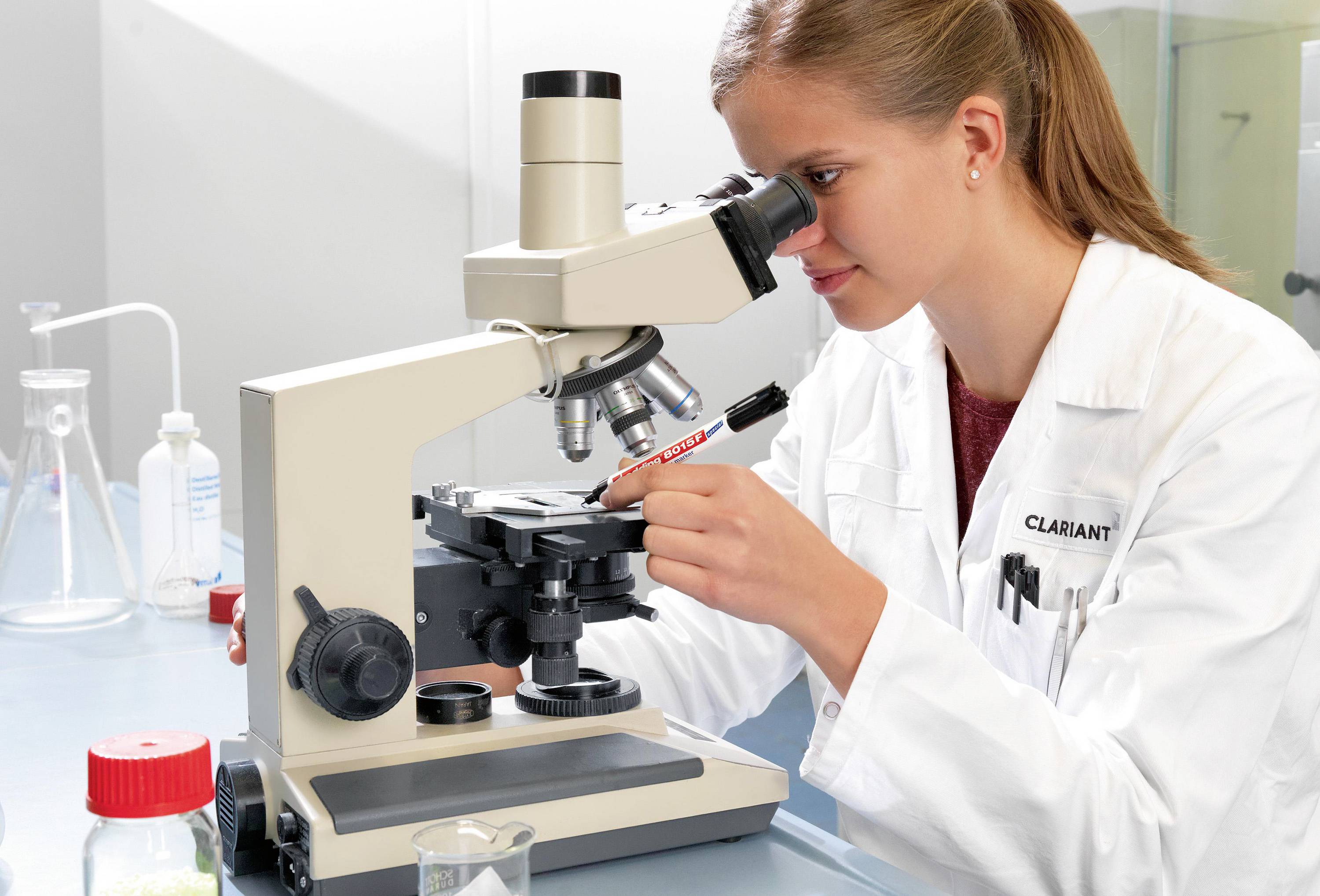 Žena v bílém plášti používá mikroskop v laboratoři. V pozadí jsou viditelné skleněné láhve a laboratorní vybavení.