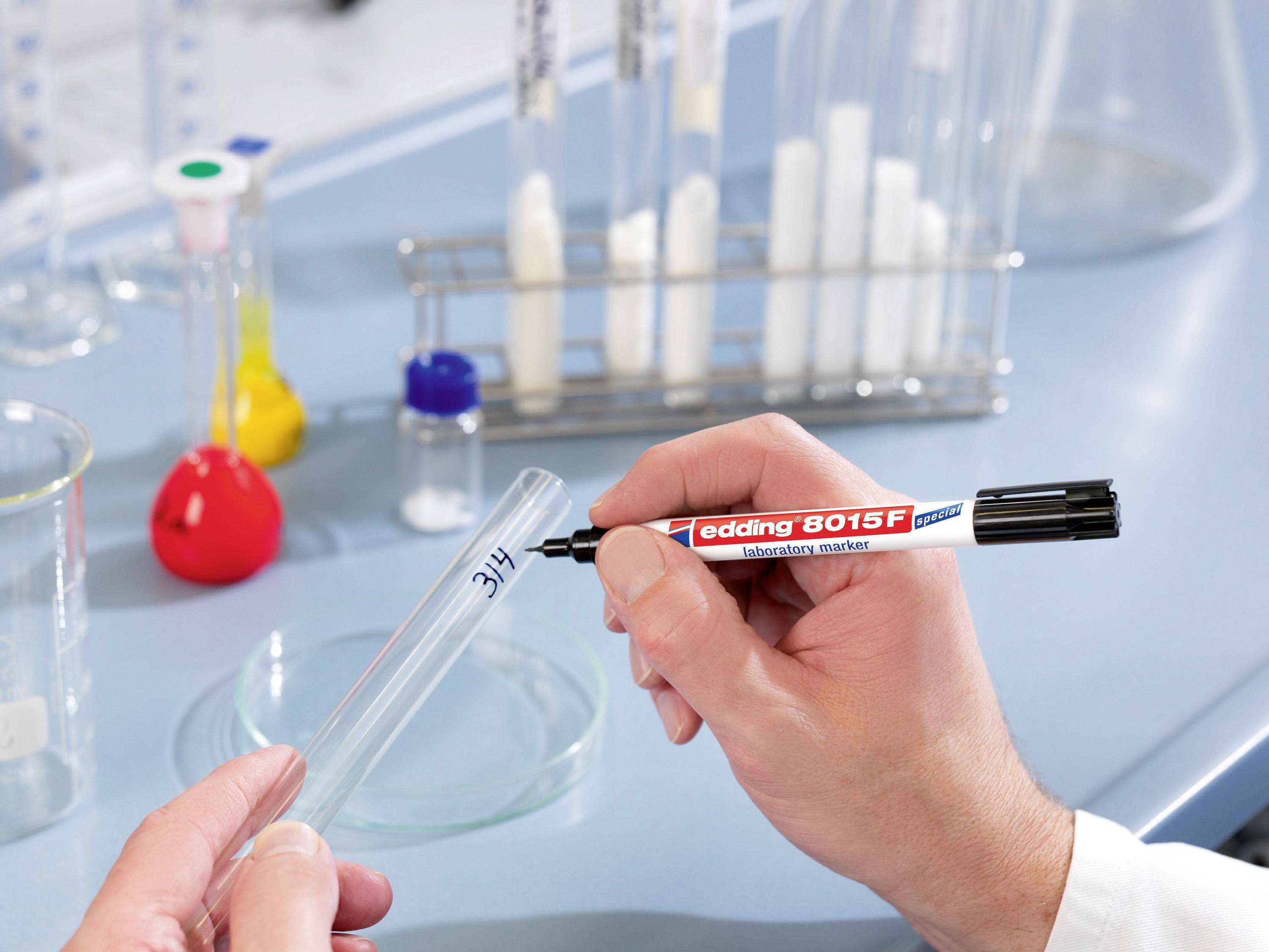 Ruka popisuje zkumavku černým fixem v laboratoři. V pozadí jsou viditelné další laboratorní pomůcky.