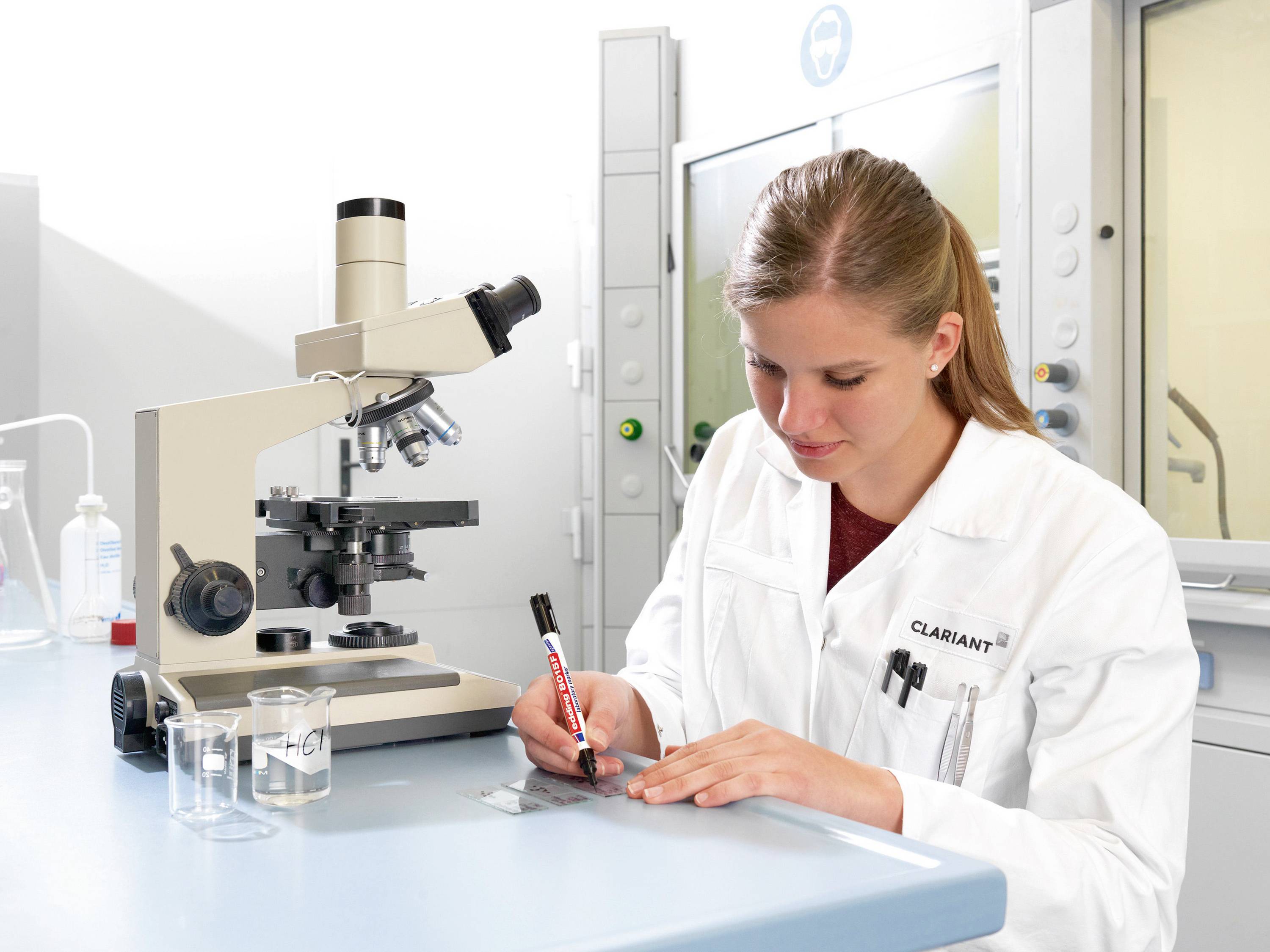 Žena v laboratoři píše na podložní sklíčko vedle mikroskopu, oblečená v laboratorním plášti. V pozadí je vidět laboratorní vybavení.