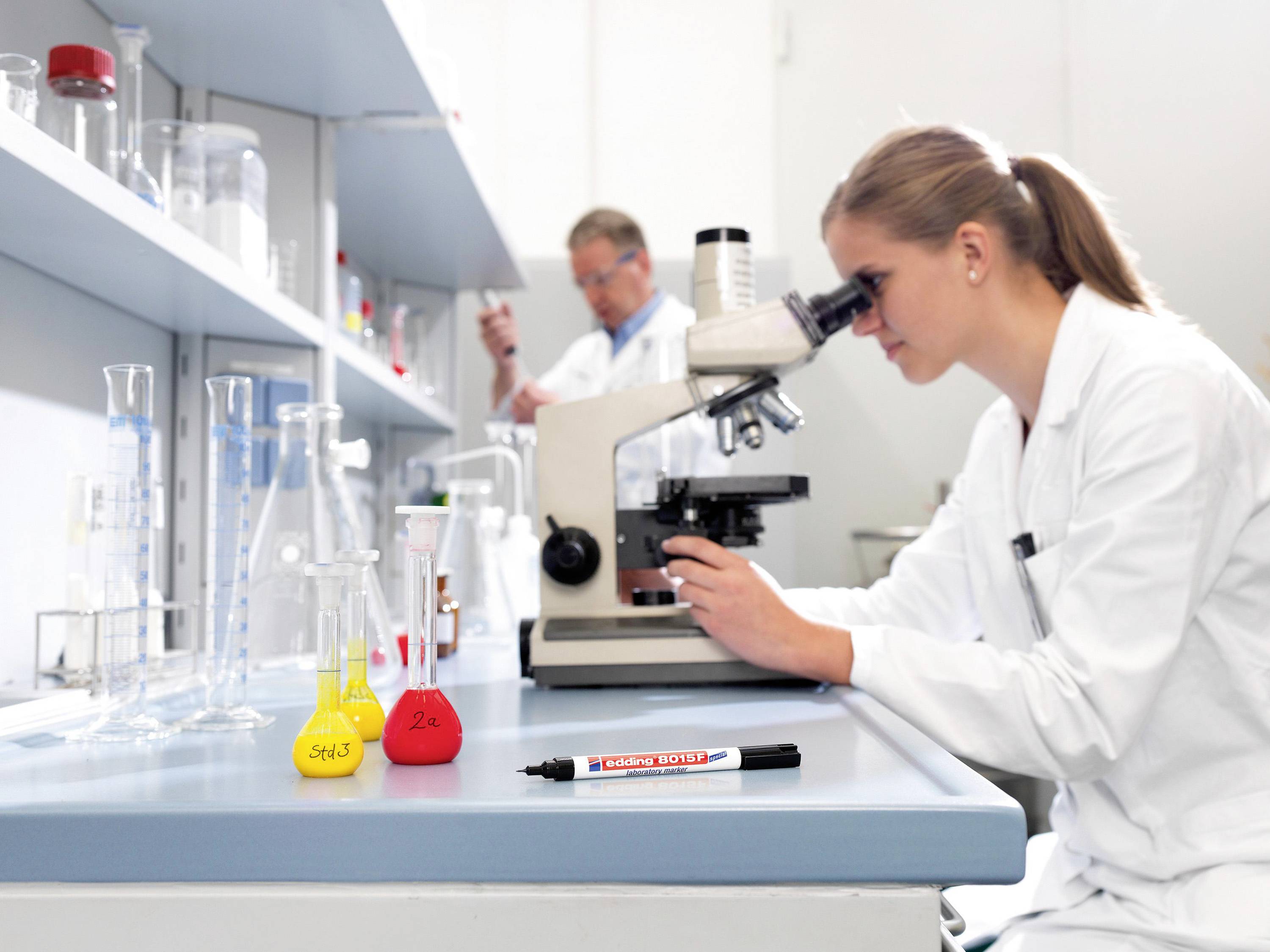 Vědkyně zkoumá preparát pod mikroskopem v laboratoři, zatímco v pozadí její kolega pracuje s chemikáliemi.