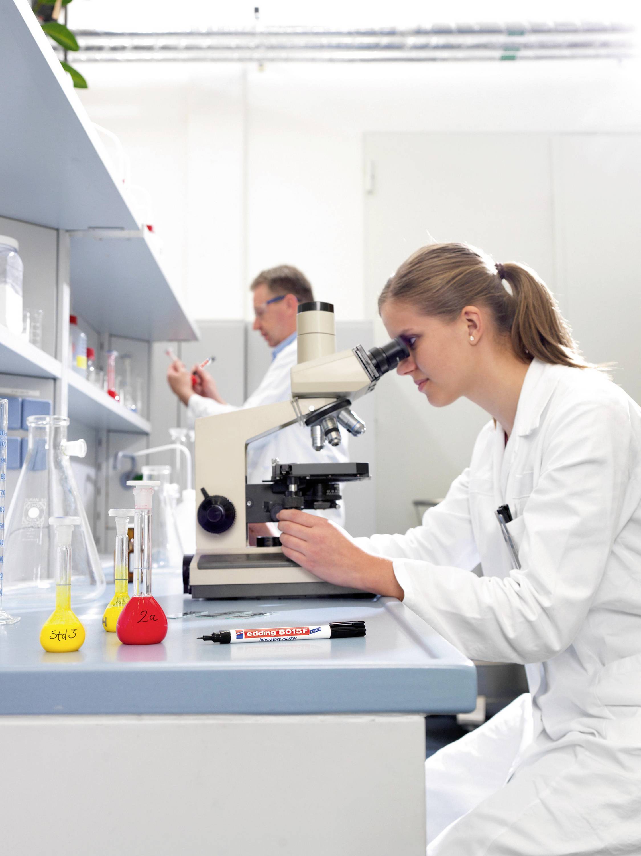 Žena v laboratoři používá mikroskop, zatímco v pozadí muž pracuje s měřicími přístroji. Na stole jsou láhve s chemikáliemi.