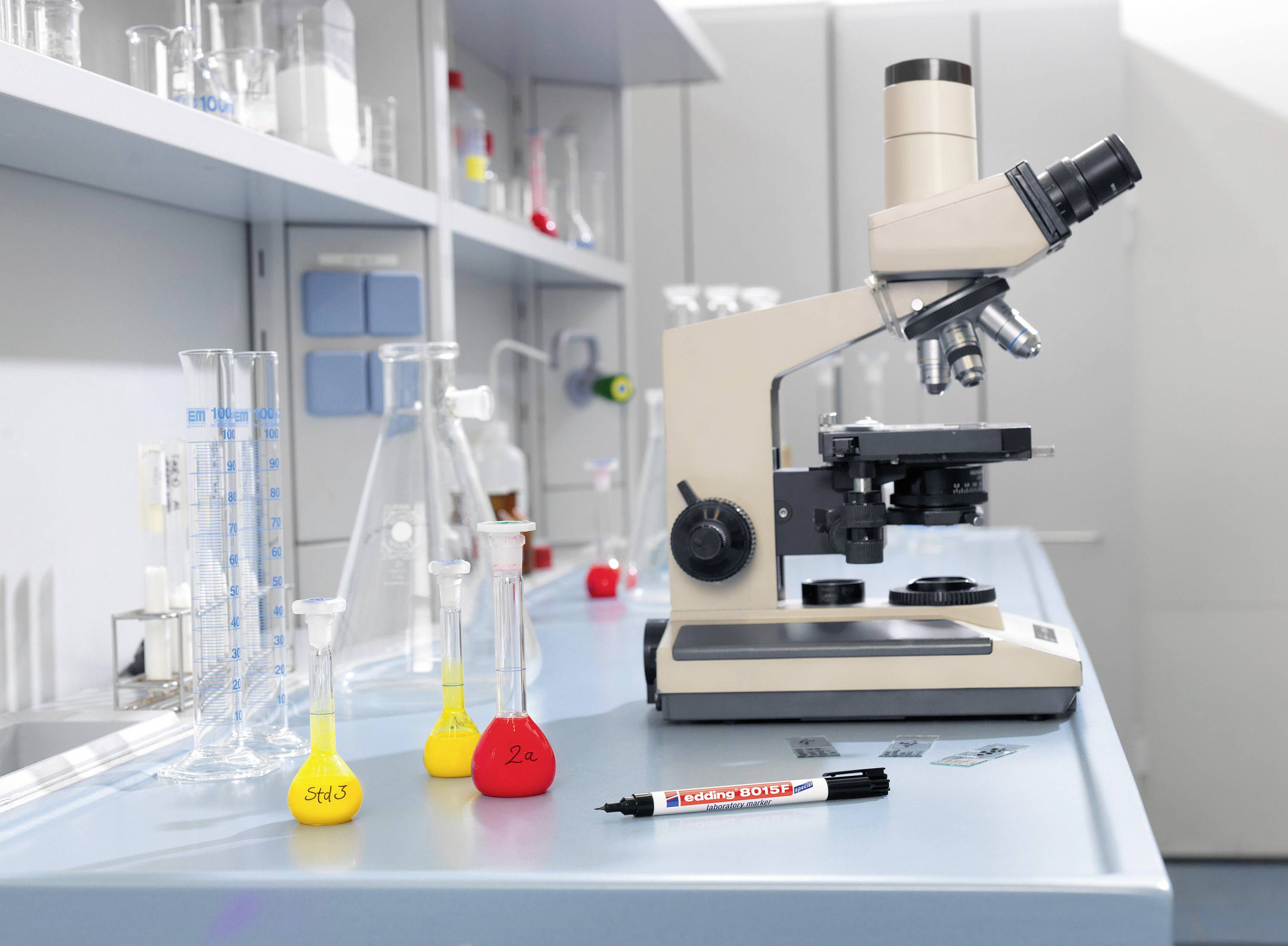 Laboratoř s mikroskopem, odměrnou baňkou a fixem na laboratorním stole. V pozadí regál s zkumavkami.