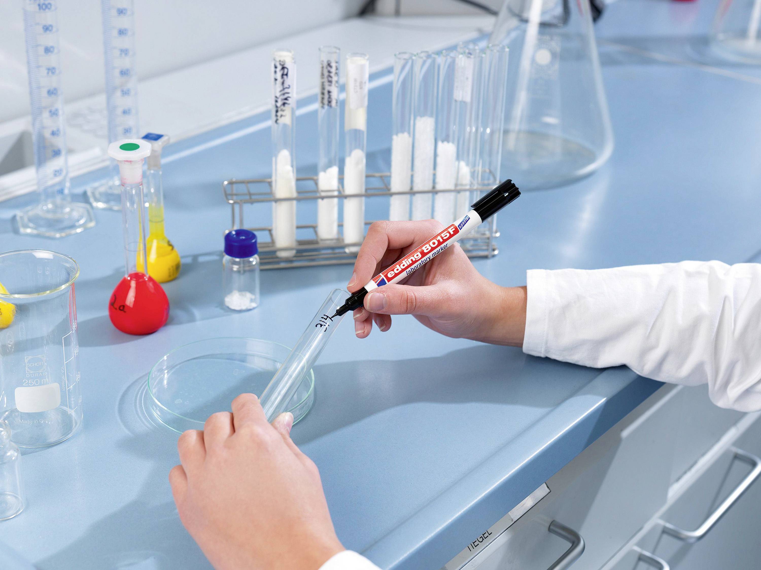 Osoba popisuje zkumavku v laboratoři, obklopená dalšími laboratorními pomůckami, jako jsou baňky a stojany na zkumavky.