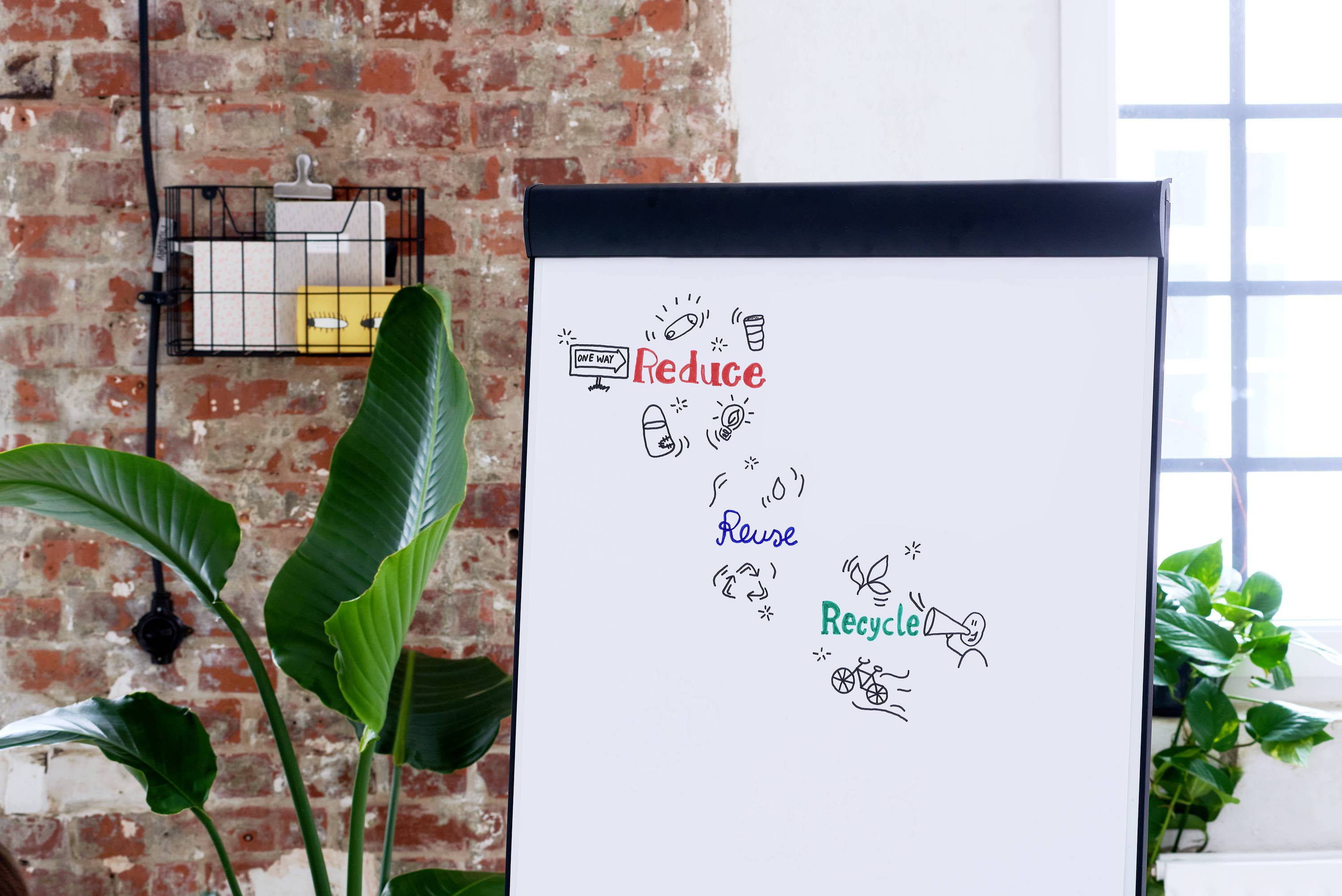 Flipchart s hesly 'Redukce, Znovupoužití, Recyklace' a odpovídajícími ilustracemi ke každému slovu stojí v světlé místnosti s rostlinami.