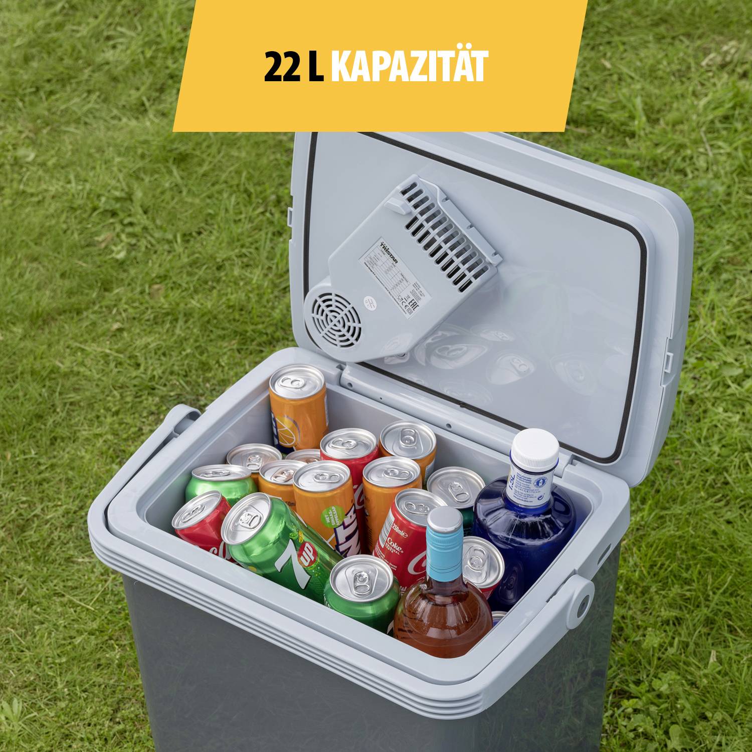 Otevřená chladicí box o objemu 22 litrů, naplněná různými plechovkami a lahvemi, stojící na travnaté ploše.