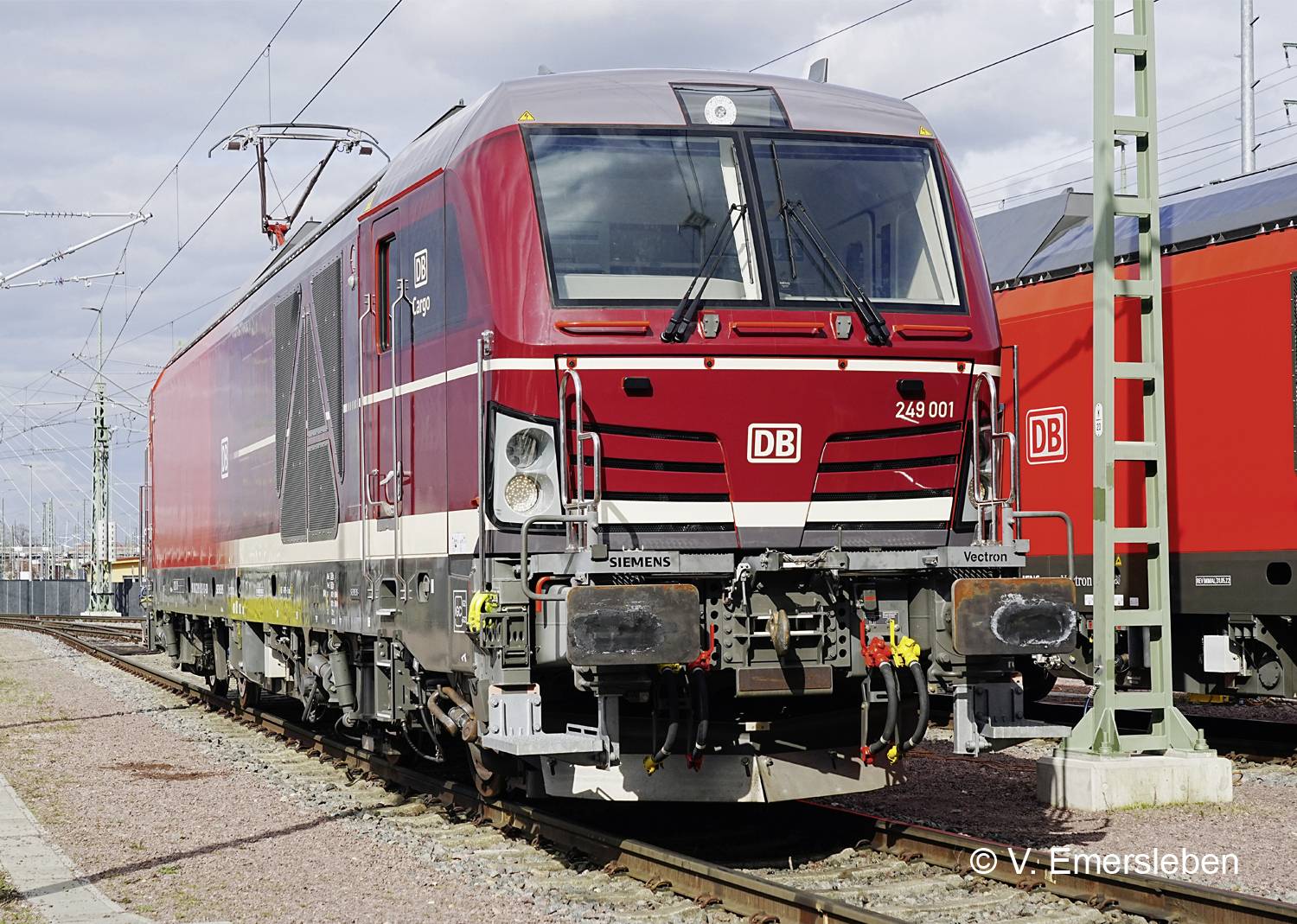 Červená a bílá lokomotiva německé železniční značky 'Siemens Vectron' stojí na kolejích. V pozadí jsou viditelné trakční vedení.