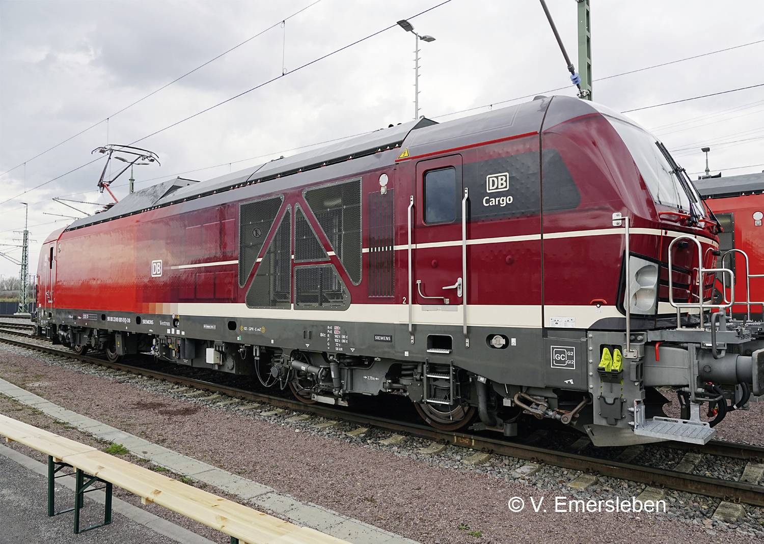 Červená nákladní lokomotiva DB Cargo na kolejích, pohled z boku, s nadzemním vedením a oblohou s mraky v pozadí.