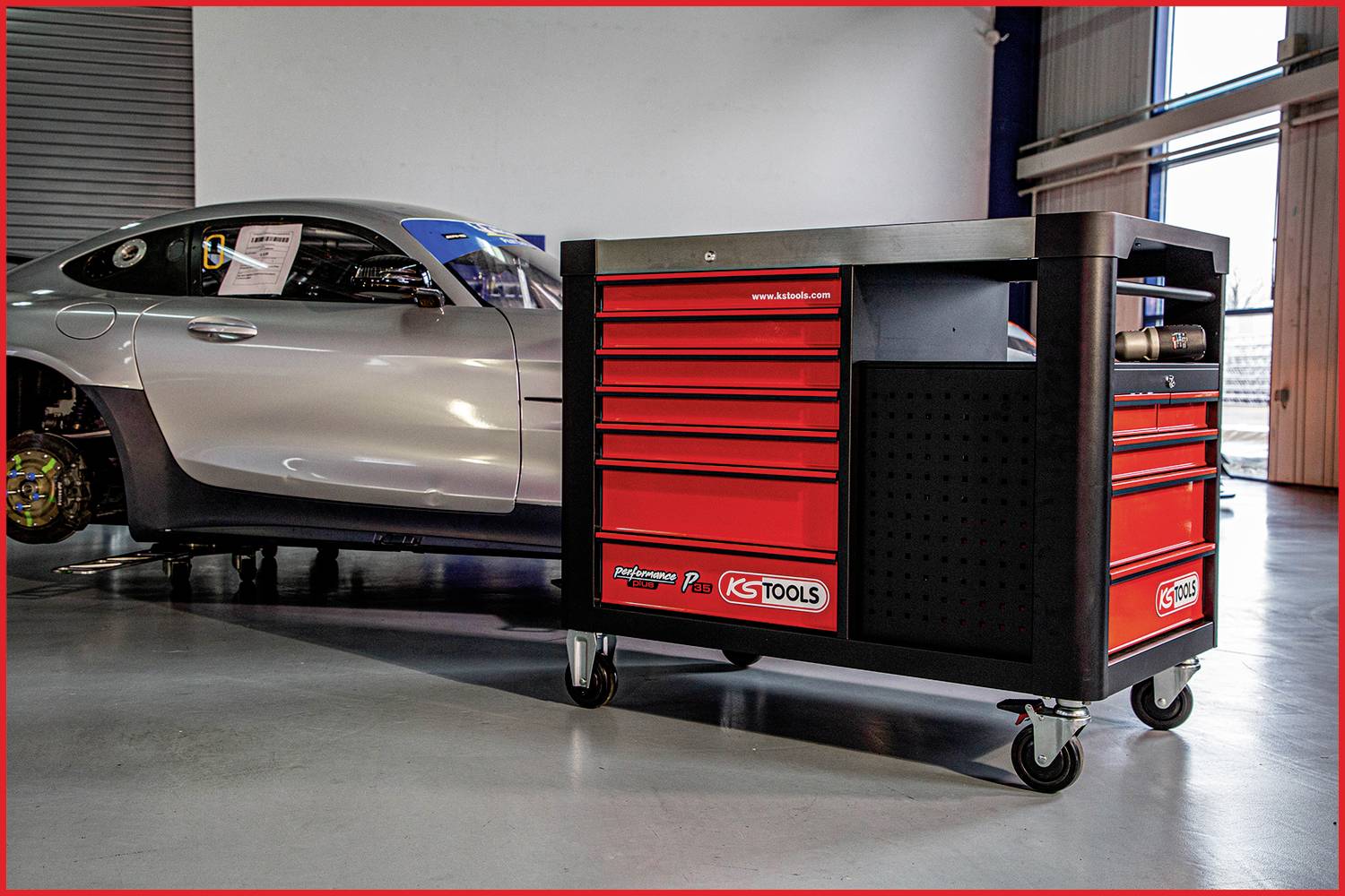 Stříbrné auto v autoservisu, umístěné vedle červeného pojízdného vozíku s nářadím, stojící na šedé podlaze.