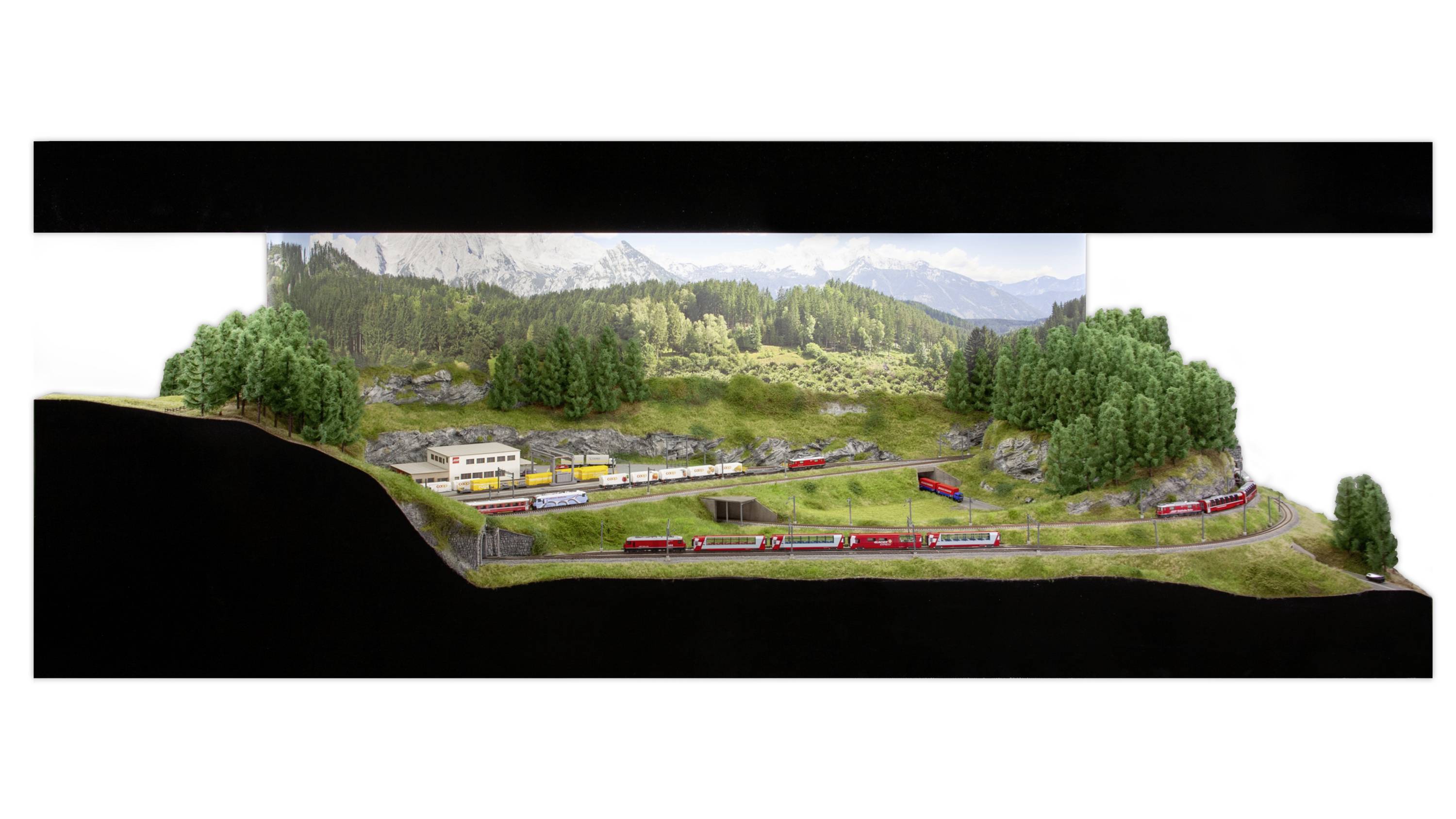 Krajinné diorama s vlaky projíždějícími zvlněnou, zalesněnou krajinou. V popředí je vidět železniční stanice.