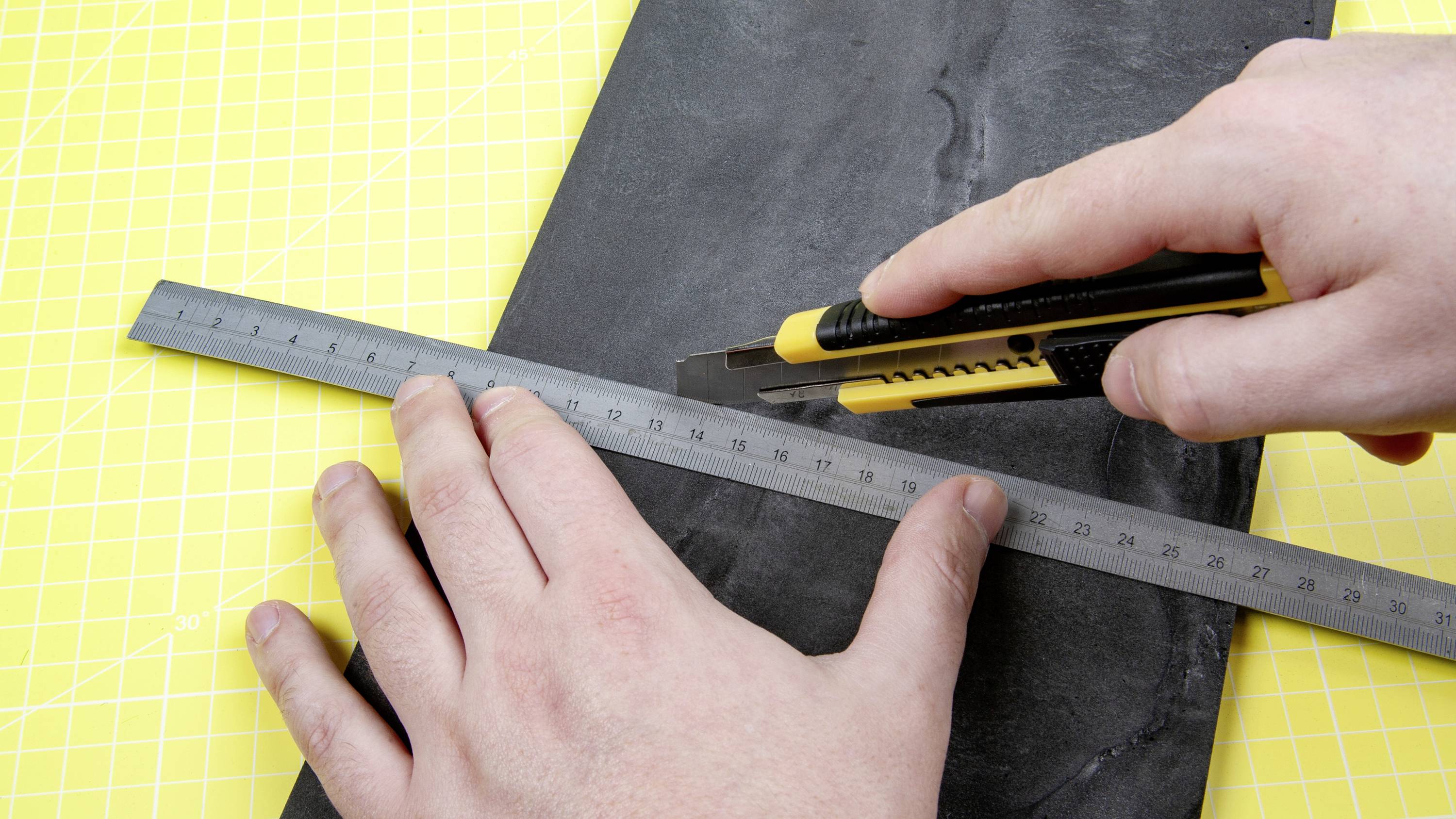 Jedna ruka drží pravítko na tmavé desce, zatímco druhá ruka vede řezací nůž. Pozadí je žluté.