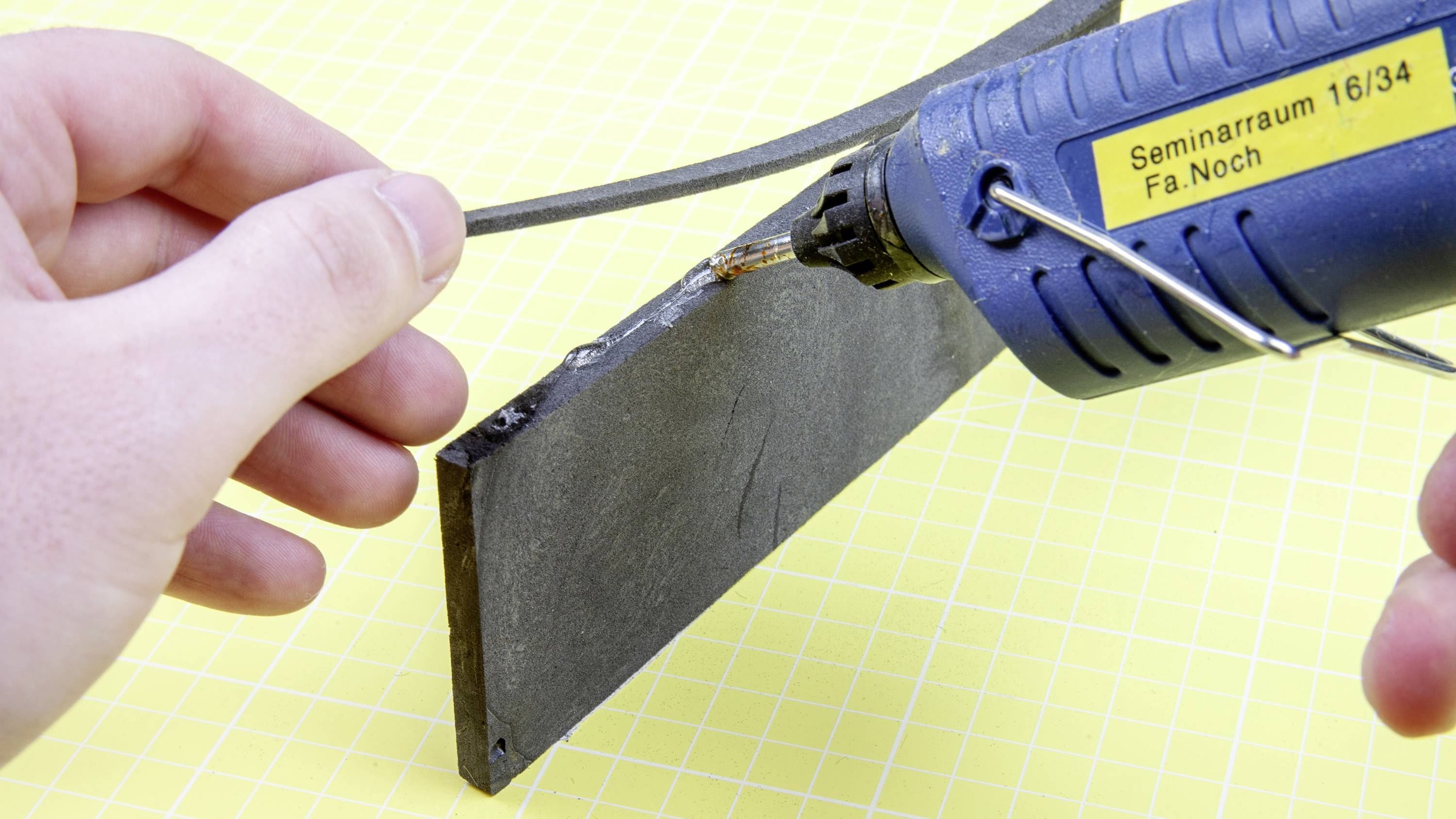 Osoba používá tavnou pistoli, aby slepila dva materiály dohromady. V pozadí je vidět žlutý kostkovaný papír.