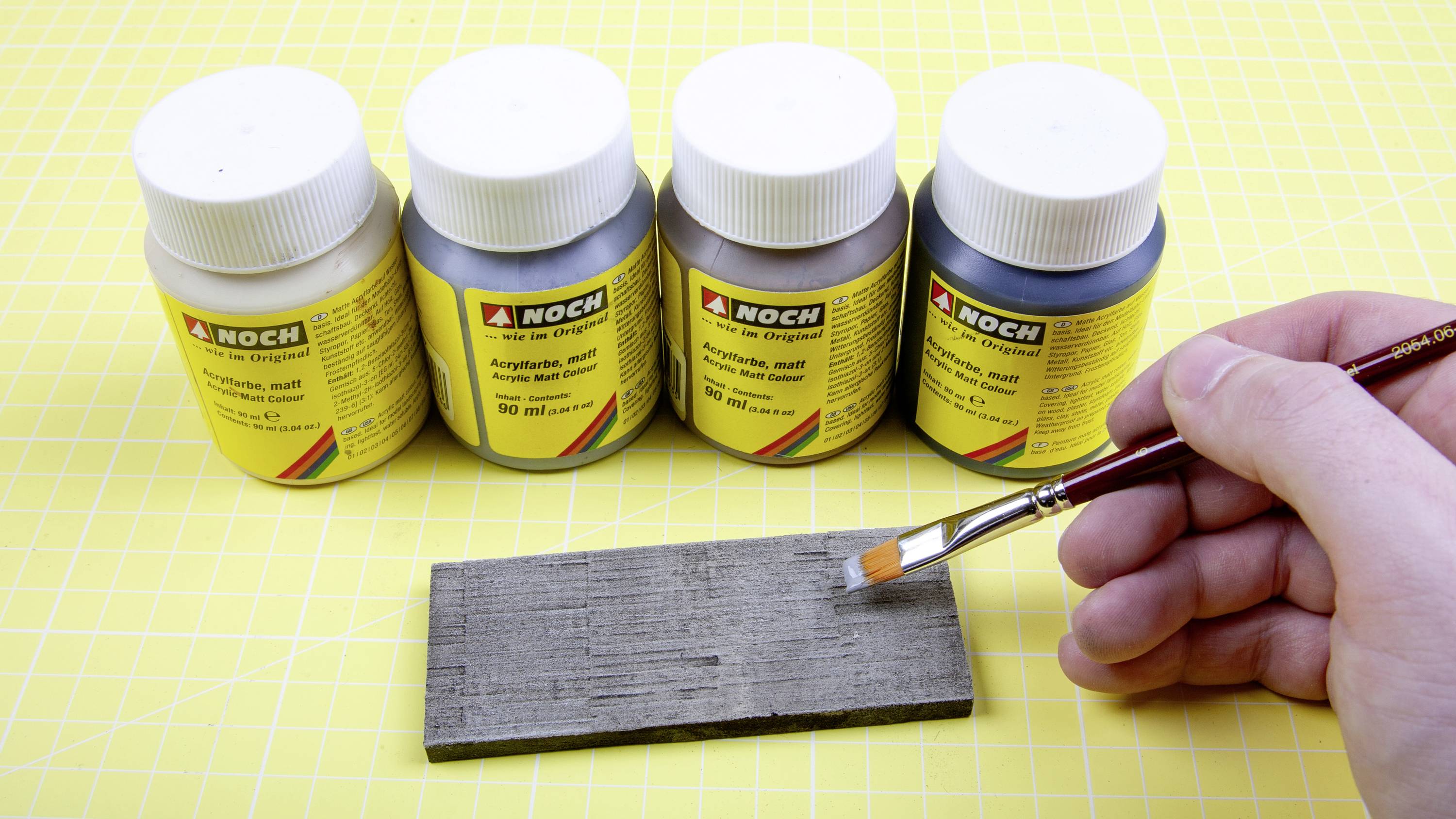 Čtyři barevné lahve na stole, každá s nápisem "Akrylová barva, matná". Ruka drží štětec a nanáší barvu na dřevěnou desku.