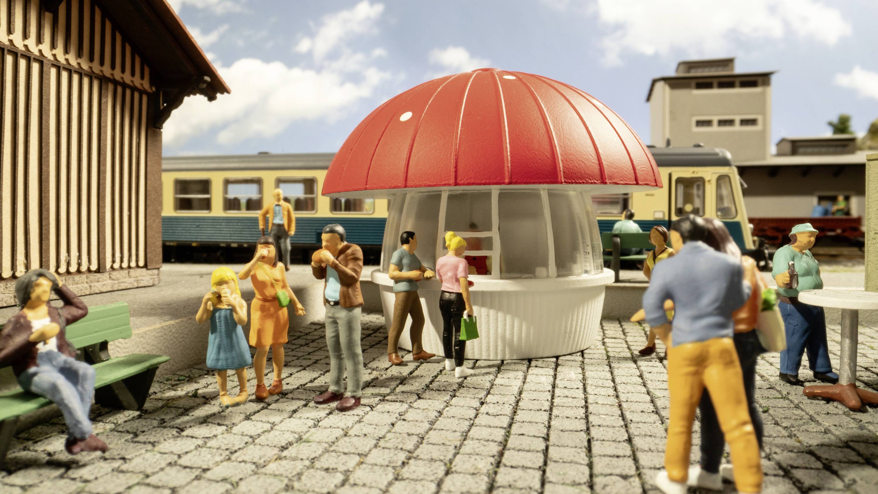 Miniaturní model nádraží s figurkami. Lidé stojí kolem červeno-bílého stánku. V pozadí jsou vidět vlaky a budova.
