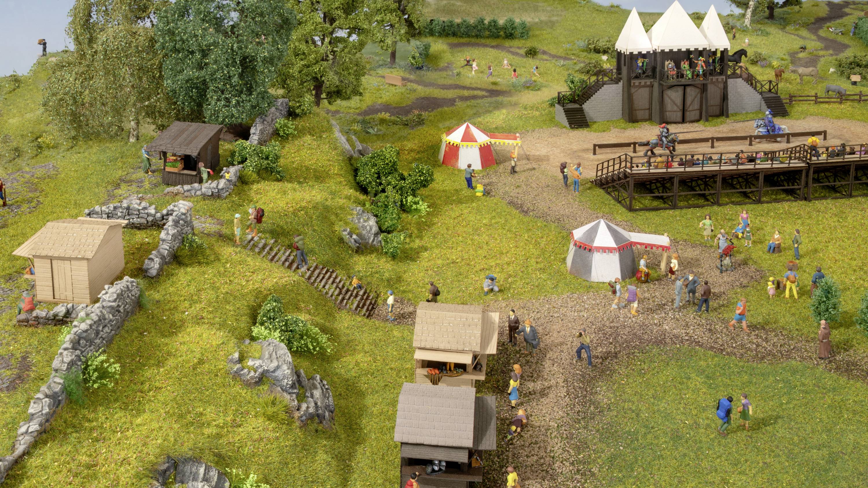 Miniaturní model středověkého trhu s stany, dřevěnými stánky a figurkami v dobovém oblečení na zelené louce.