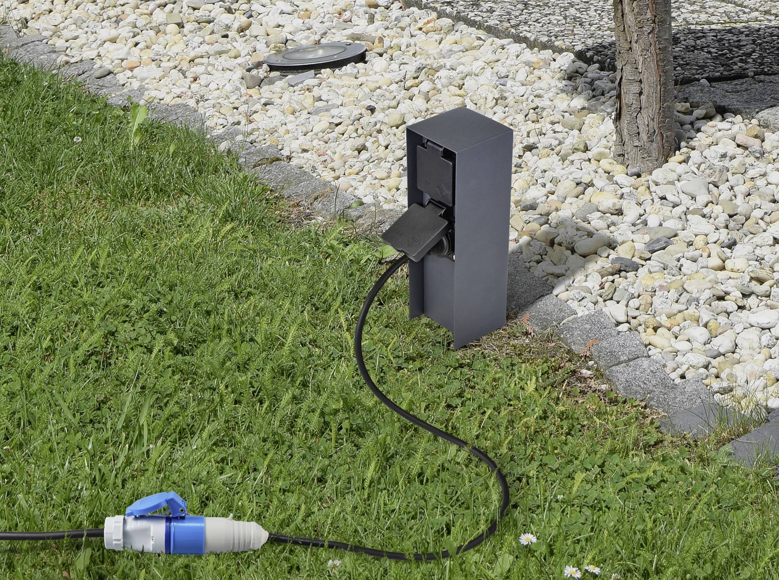 Černá venkovní zásuvka na trávníku s připojeným kabelem, obklopená oblázky a se stromem v pozadí.