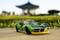 Zelené hračkové auto v popředí stojí na betonové ploše. V pozadí je rozmazaný pavilon.